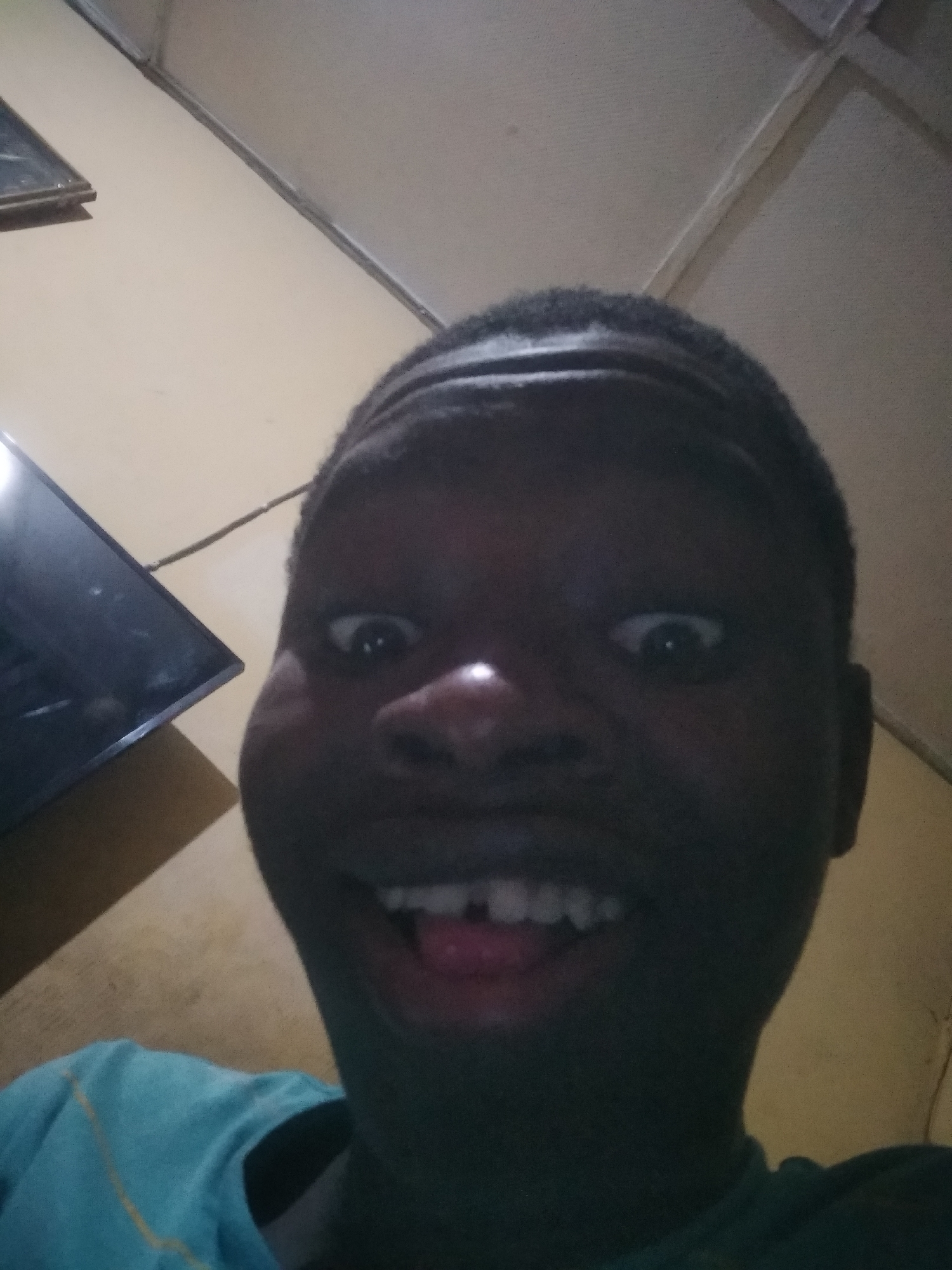 A person is taking a selfie. The person is facing the camera. The person is smiling and appears to be in their home in Shangisha, Nigeria. A television is visible on the wall in the background. The subject is wearing a blue t-shirt.