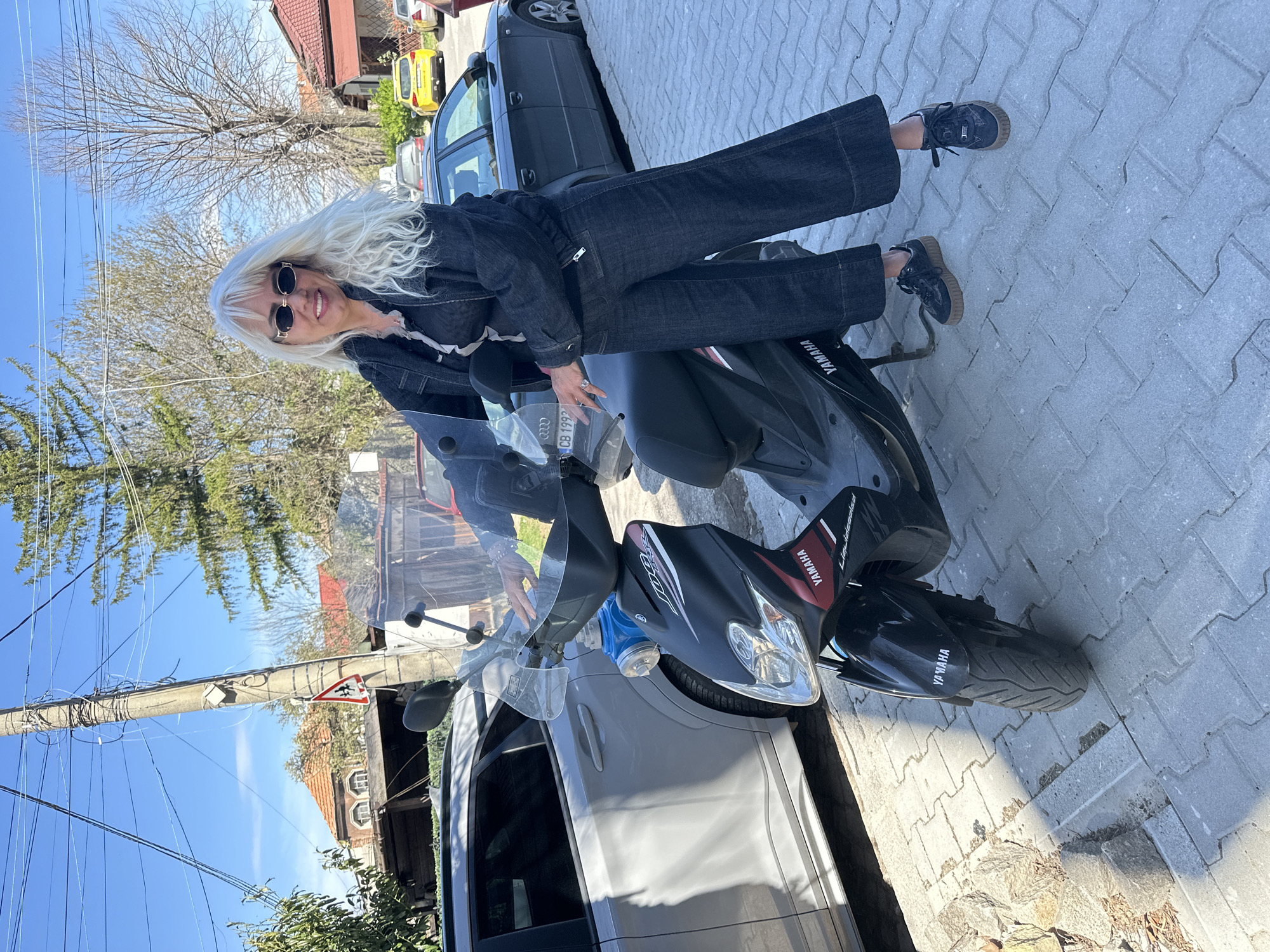 A woman with long, platinum blonde hair and sunglasses is sitting on a black Yamaha scooter in front of a silver car. She is wearing a dark denim jacket and wide-legged denim pants, along with dark shoes. The scooter has a windshield and a blue container attached to the front. The background includes a grey car, a yellow car, trees with bare branches, and buildings. The ground is paved with interlocking pavers. The license plate on the scooter reads "CB 1992". The photograph appears to be taken on a street in Sofia, Bulgaria.