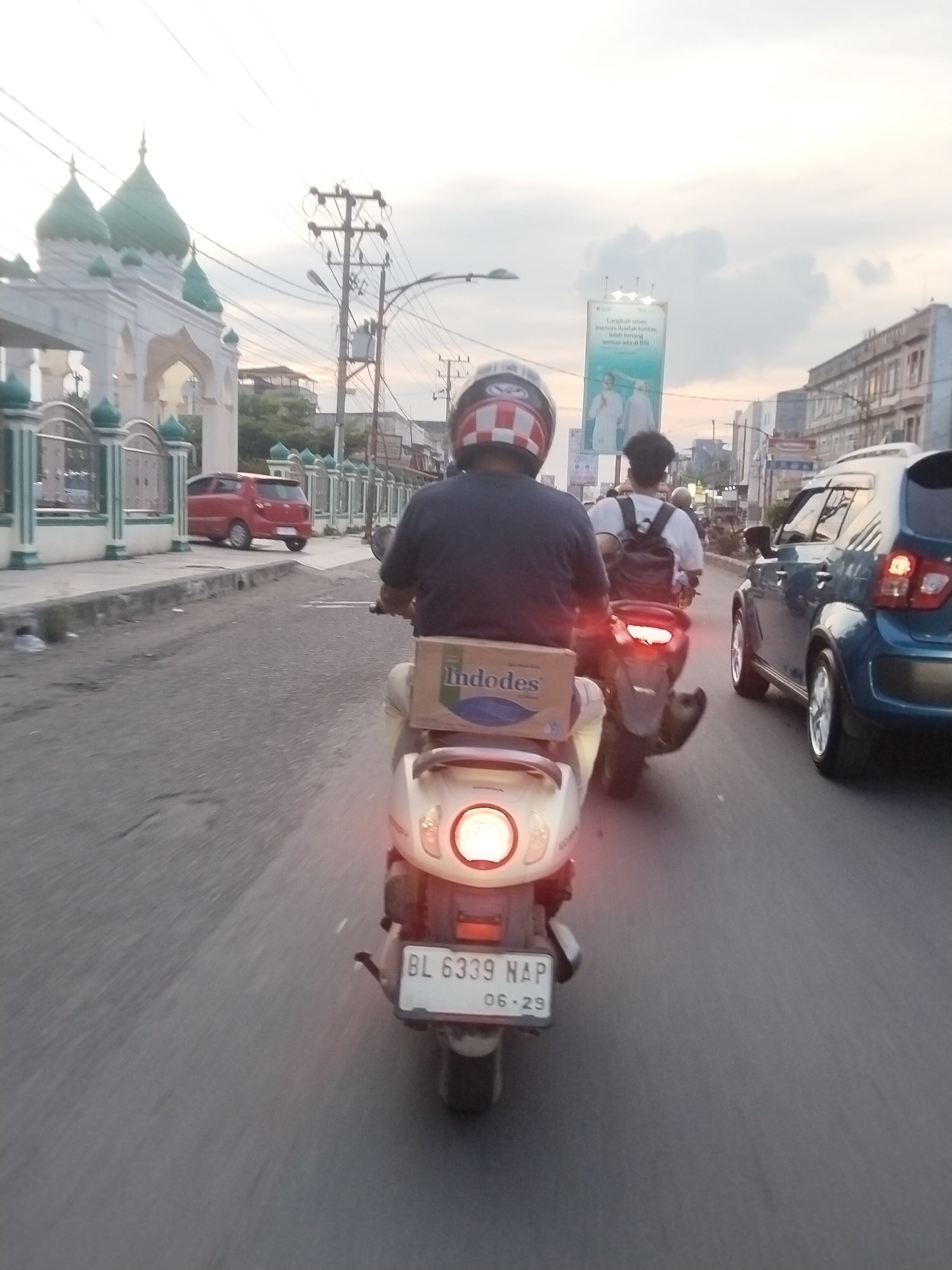 The image depicts a street scene in Lhokseumawe, Indonesia, during the late afternoon or early evening. The sky is overcast with pale clouds.
In the foreground, a person wearing a red, white, and blue striped helmet is riding a light-colored scooter. The scooter has a license plate that reads "BL 6339 NAP 06.29". A brown cardboard box labeled "Indofood" is strapped to the back of the scooter. The scooter's taillight is illuminated.
Behind the first scooter, another scooter is visible, ridden by a person wearing a white t-shirt and a backpack. The second scooter has a red taillight.
Further ahead, a blue car is driving in the same direction as the scooters. To the left of the road, a red hatchback car is parked.
The backdrop of the scene includes buildings, utility poles with numerous wires, and a mosque with green domes and white architecture. A billboard with an advertisement featuring two individuals is also visible. The road surface appears to be asphalt. The overall impression is a typical urban street environment with traffic and everyday activities.