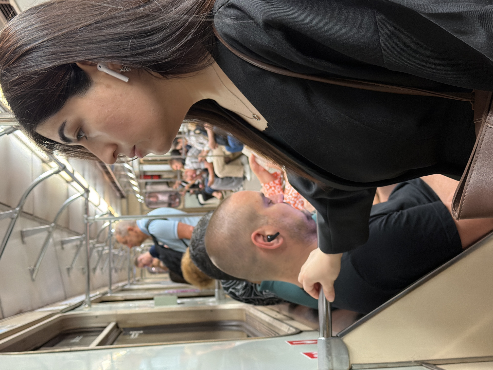 The image shows a woman with dark hair, wearing a black blazer and a necklace, riding on a Moscow Metro train. She has a white earbud in her right ear. In the foreground, a man with a shaved head, wearing a black t-shirt and a black earbud, is seated next to her. His hand is resting on a metal pole. Other passengers are visible in the background, some seated and some standing. The interior of the train car appears worn, with light-colored seats and metal overhead railings. The scene is set inside a moving train.
