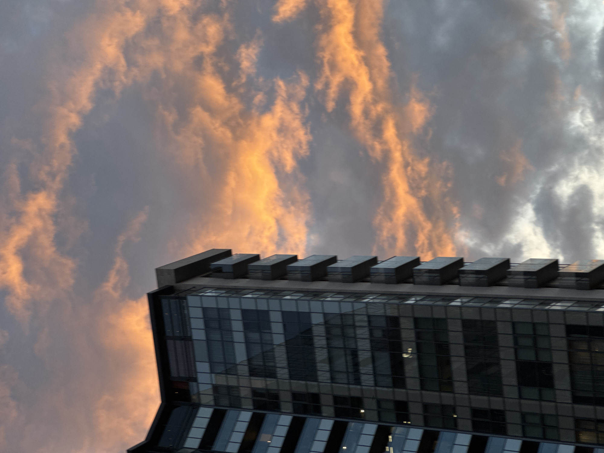 A low-angle shot depicts the top of a modern building against a dramatic sky, which is indicative of dusk or dawn. The sky is filled with clouds that transition from grey to hues of orange and peach, suggesting the presence of sunlight reflecting off the clouds. The building has a multi-tiered, rectangular design with a series of small protruding structures at the top. The facade is predominantly glass, arranged in a grid-like pattern, reflecting the sky and hinting at a cityscape. The overall composition is characterized by a contrast between the architectural design and the natural atmospheric display. Yerevan, Armenia is not explicitly identified within the visual frame.
