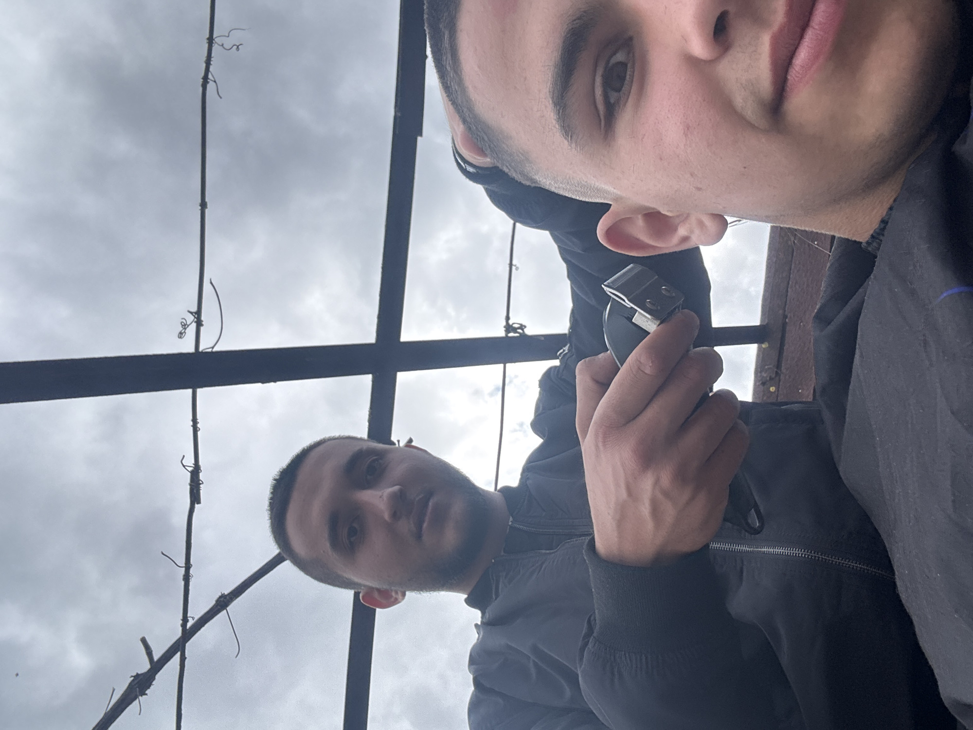 The photograph shows two young men in an outdoor setting under an overcast sky. The framing is unusual, tilting upwards as if the camera is positioned low. One man, positioned at the upper right of the image, is holding a hair clipper near his ear. The second man is visible in the lower left, looking towards the camera. Both are wearing dark-colored jackets. The location appears to be under a metal structure with vertical and horizontal beams. Thin vines, likely dead, are clinging to these beams. The description includes no information to prove that the location is Topoli, Bulgaria.