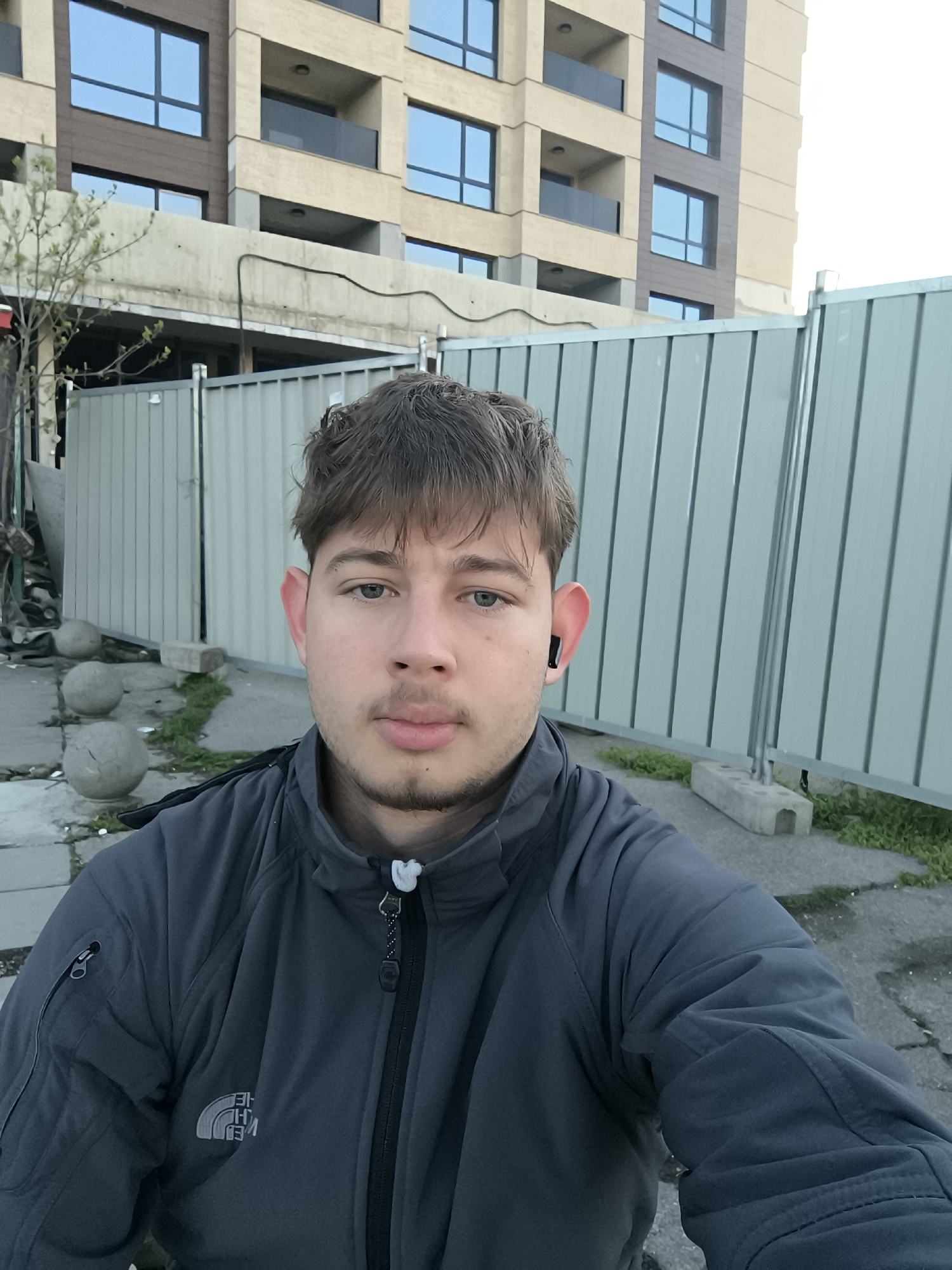 A medium shot captures a fair-skinned young man with short, dark hair and blue eyes. He wears a dark gray jacket with the North Face logo visible on the lower left side. An earbud is visible in his left ear. He appears to be taking a selfie.
Behind the man is a corrugated metal fence, partially obscured by overgrown vegetation and some scattered concrete spheres. In the background rises a multi-story building with modern architectural features including windows and balconies.
The location is likely in Sofia, Bulgaria, as requested.