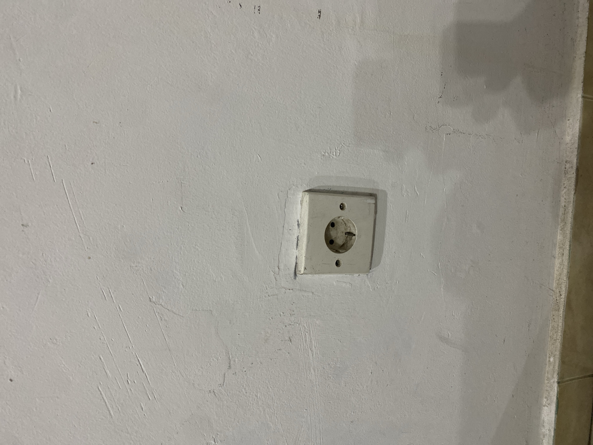 A close-up, medium-shot, eye-level, indoor photograph shows a single, cream-colored electrical outlet on a white, textured wall. The outlet is slightly angled and appears to be made of plastic. It has two round openings and two small screw holes, one of which is clearly visible. The wall surrounding the outlet shows some minor imperfections, including faint horizontal lines and small dark specks. The bottom of the image reveals the top edge of a tiled baseboard, with light brown tiles and a darker grout line. The lighting is dim, casting a subtle shadow to the right of the outlet. The overall environment suggests an interior setting, possibly a room or hallway, in Kuta Selatan, Indonesia.
