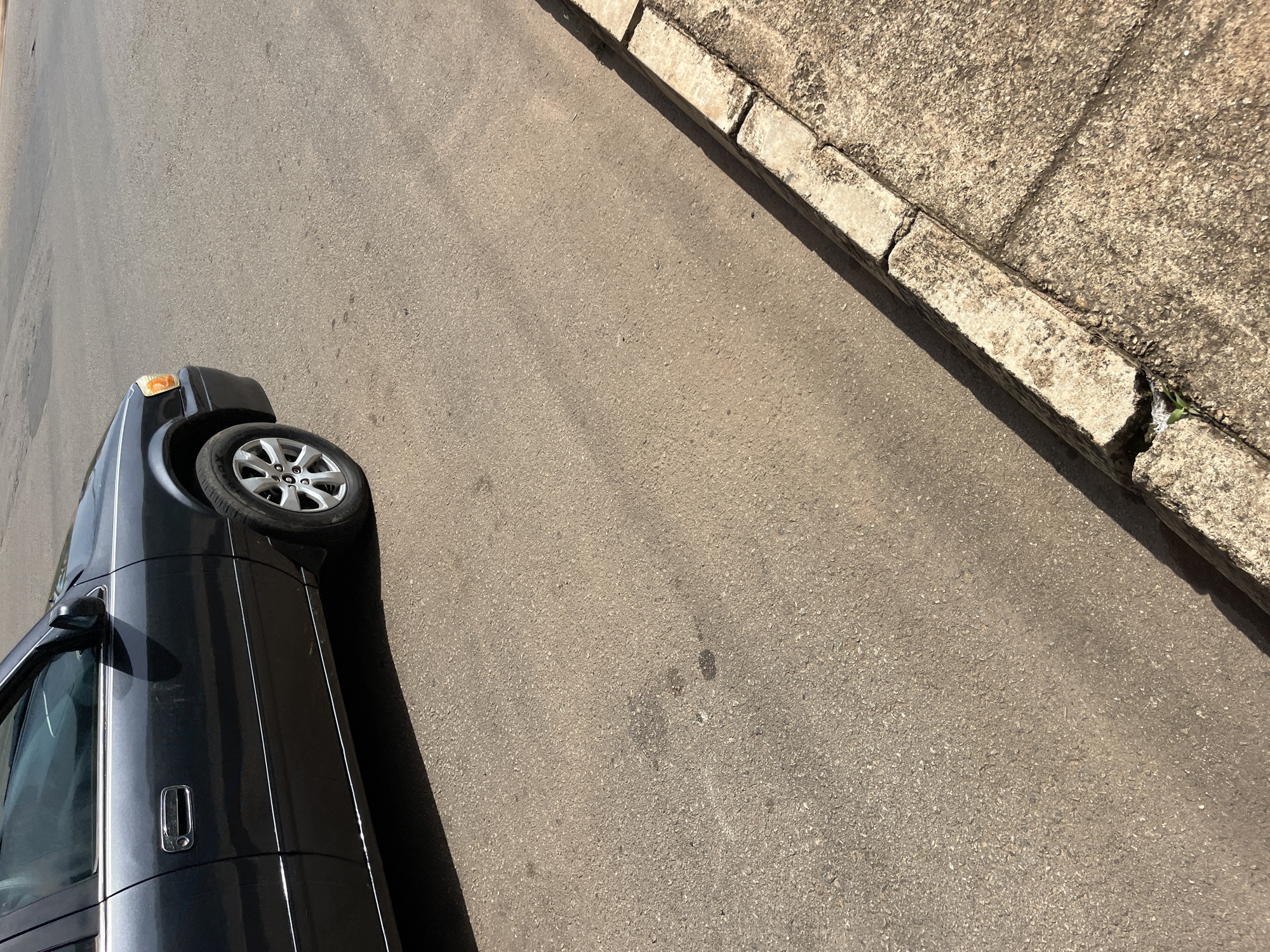 The image shows the side view of a dark-colored car parked on an asphalt road. The car is positioned diagonally, with its front end facing towards the upper left of the frame. The car's wheel is visible, as is part of its body, including the door handle and side mirror.
Adjacent to the road is a concrete curb and sidewalk. The curb appears weathered and cracked in places, with a small green plant growing out of a crack near the bottom right of the frame. The asphalt road itself is a uniform gray color, with some faint darker markings and specks of lighter aggregate visible. Sunlight casts a shadow of the car onto the road, indicating it is daytime. The overall scene suggests an urban or suburban setting in Bukuru, Nigeria. There is no text visible in the image.