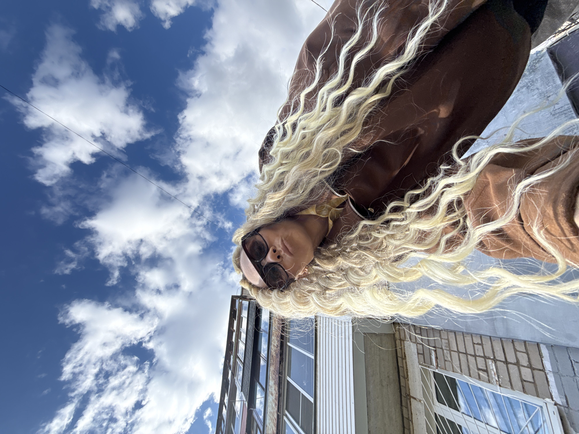 The image captures a unique low-angle perspective of a person with striking, long, wavy, blonde hair and dark sunglasses. The subject is seen from below, with their head positioned towards the bottom-left of the frame and their body and hair extending diagonally upwards and to the right, creating a sense of dynamic movement against the sky. They are wearing a brown jacket.
The background is dominated by a clear blue sky interspersed with scattered, fluffy white clouds, suggesting a bright, daytime setting. A thin, dark power line stretches across the upper left portion of the sky. To the bottom right, parts of urban buildings are visible, characterized by multiple windows reflecting the sky, brickwork, and light-colored paneling, indicative of a developed area. The overall composition is disorienting yet artistic due to the extreme upward angle and the subject's diagonal orientation. There is no visible text in the image. The location context for this scene is Rusanovo, Russia.