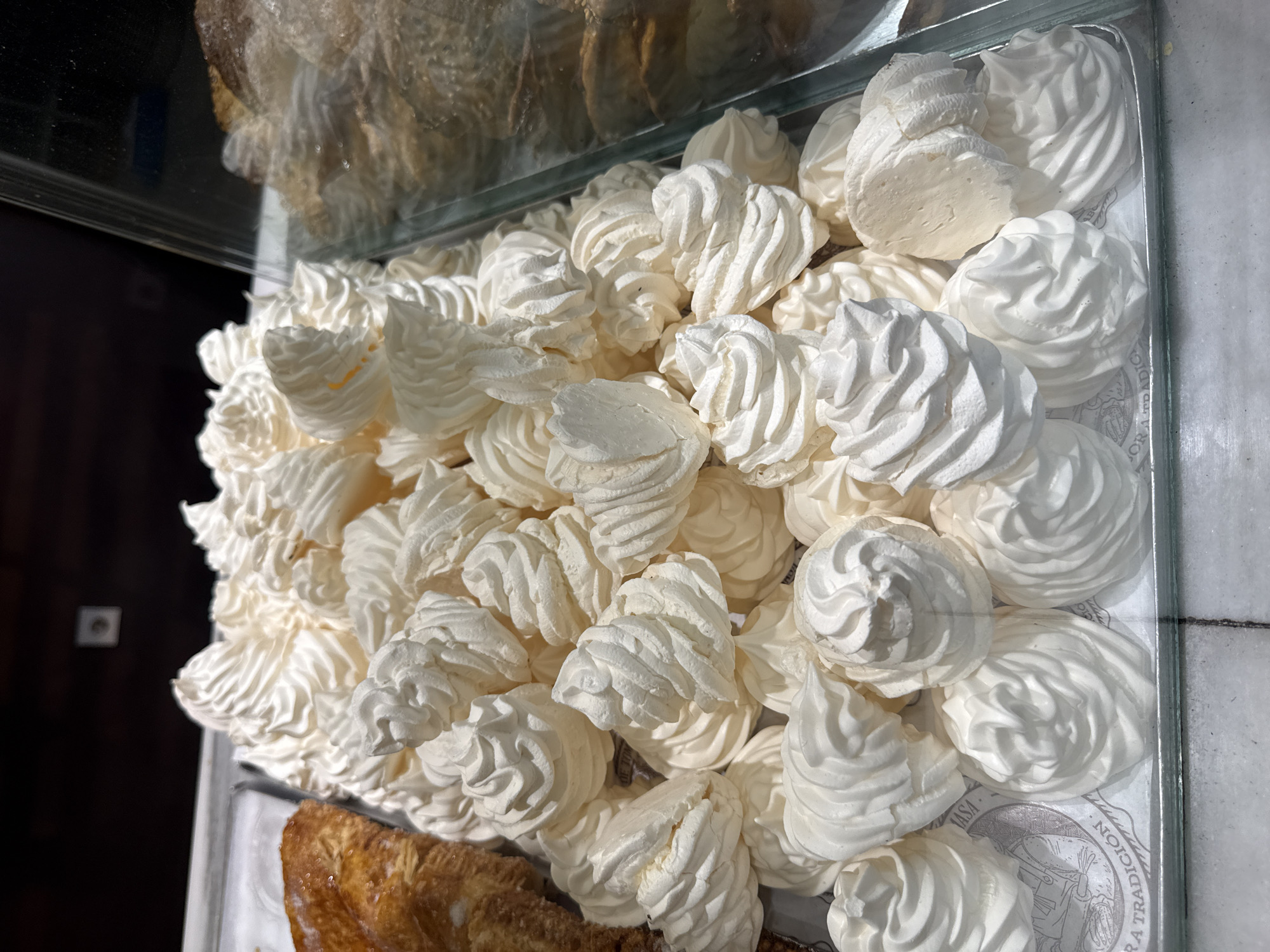A high-angle, close-up shot captures a substantial quantity of white meringues displayed within a retail setting. Approximately 50 to 70 individual meringues, predominantly piped into distinct swirled or star-tip shapes, are piled on a rectangular metallic tray. The meringues exhibit a matte, crispy texture; some pieces show minor fragmentation or irregular forms. A faint yellow discoloration is visible on select meringue pieces. The tray is lined with white paper featuring a subtle grey pattern, partially revealing text such as "RA TRADICION" and "ASA" at the bottom left.
To the immediate left of the meringues, a portion of a golden-brown, flaky pastry is visible on an adjacent tray. A clear glass or acrylic barrier runs along the right side of the meringue display, separating it from an obscured area containing blurry, darker baked goods. The glass reflects parts of the dark background. The upper background is dark, with a small, light-colored square object, possibly an electrical fixture, visible centrally. The display is situated on a light-colored surface, possibly a countertop, showing a dark seam or grout line. The location is in the city of Unknown, Unknown.