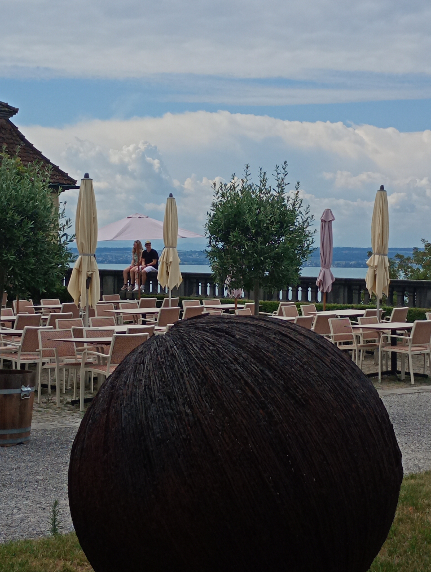 The image shows an outdoor seating area with tables and chairs, likely a cafe or restaurant, situated in Meersburg, Germany. In the foreground, a large, dark, textured sphere is prominent. Beyond the seating area, a stone balustrade overlooks a body of water, possibly Lake Constance, with hills in the distance under a cloudy sky. Two people, a man and a woman, are seated on the balustrade, facing the water. Several closed patio umbrellas are visible among the tables. A wooden barrel is positioned to the left of the seating area. The architecture of a building with a red-tiled roof is partially visible on the far left, suggesting it's part of a historical site or complex. The overall scene depicts a tranquil outdoor setting with a view of the lake.