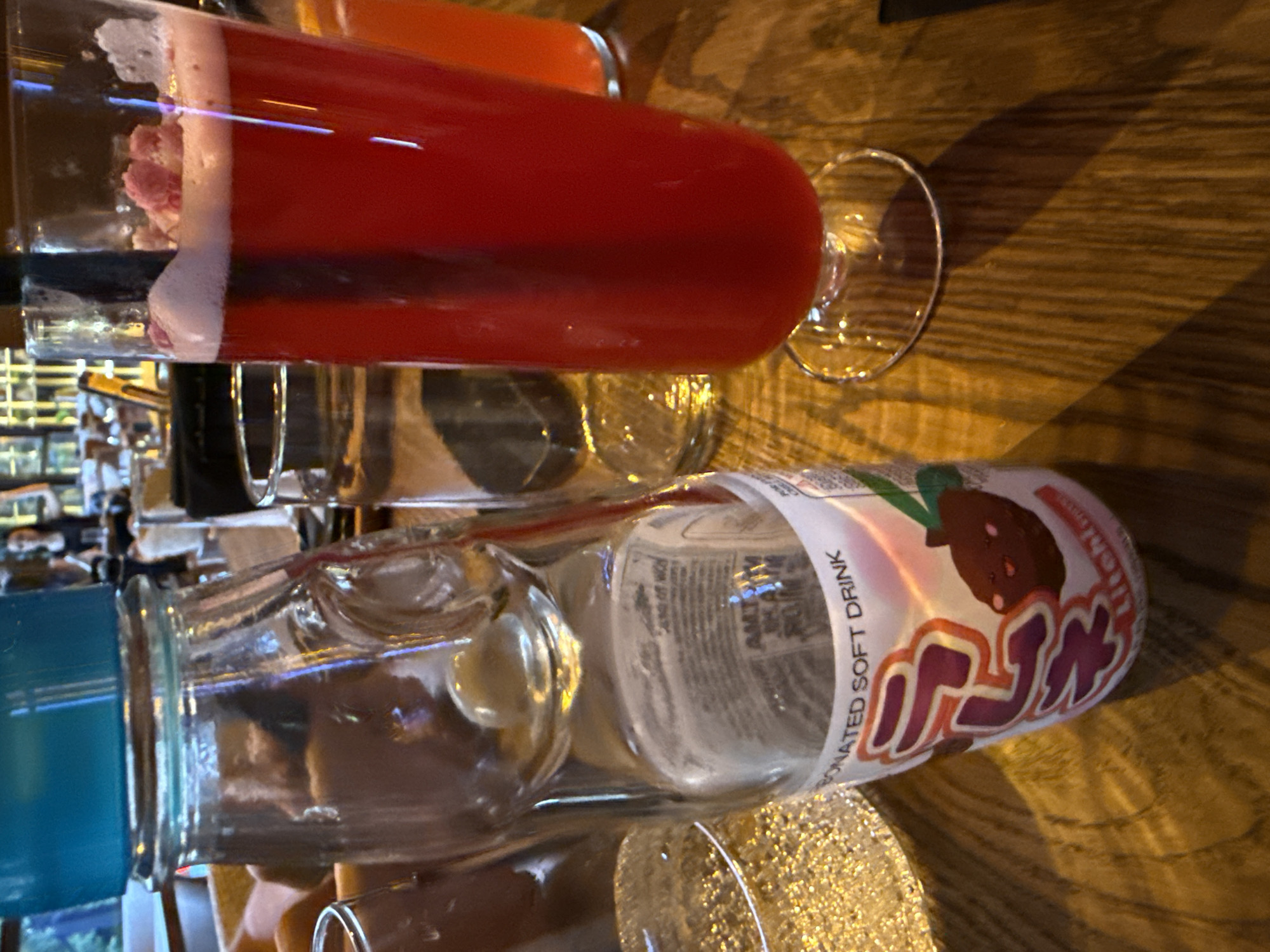 An indoor photograph shows a close-up of a wooden table with beverages. In the foreground, a clear glass bottle with a blue cap, containing a light-colored liquid and a marble stopper, sits on the left. A label on the bottle reads "CARBONATED SOFT DRINK" and features Japanese characters and an illustration of a strawberry. To the right of the bottle is a tall, clear glass filled with a bright red beverage topped with foam and small pink pieces, possibly edible flowers or ice. A dark straw extends from the glass. In the background, out of focus, other glasses and people are visible. The setting appears to be a restaurant or bar in Sofia, Bulgaria, given the overall context provided.