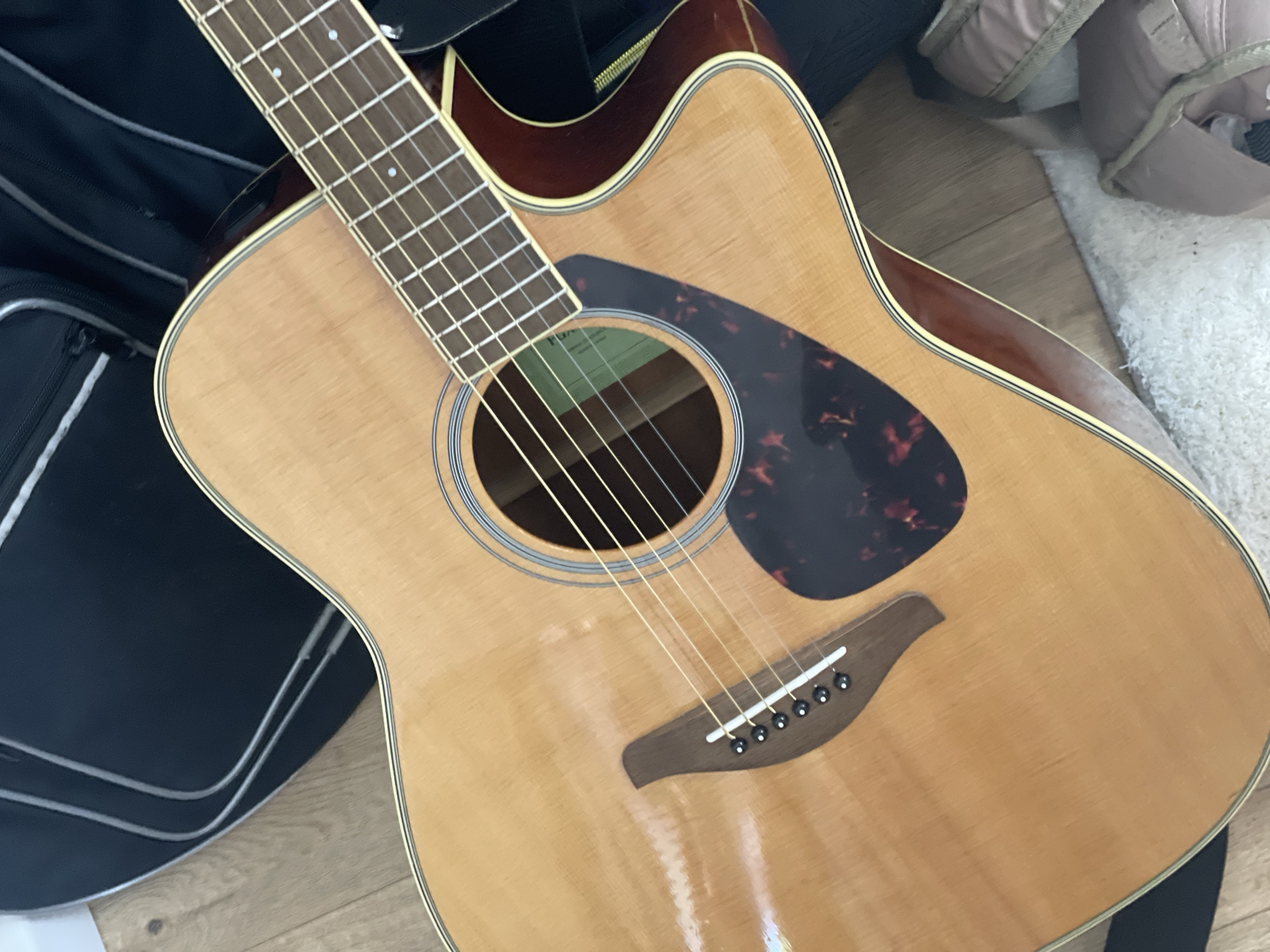 The image shows an acoustic guitar with a light-colored wood top and darker wood sides. It is resting on a wooden floor and a white rug. A black guitar case is visible in the background. The guitar has six strings, a tortoiseshell pickguard, and a cutaway body. The lighting is natural and appears to be coming from the side, casting shadows on the guitar. There are no people visible in the image. The scene is indoors and appears to be a residential setting. There is no information in the image that can be used to determine its location in the city of Ulmen, Germany.