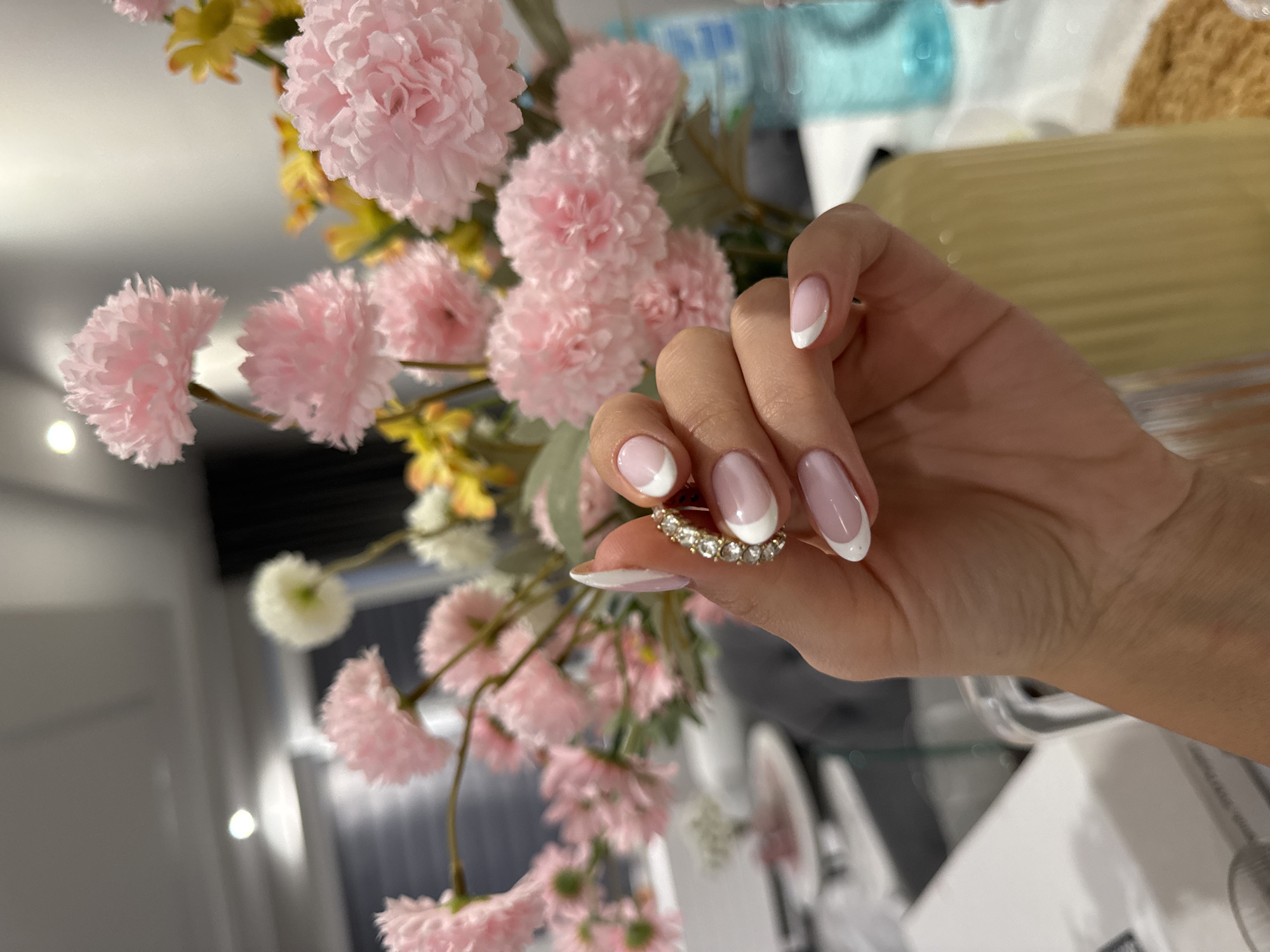 The image shows a hand holding a diamond ring. The hand has manicured nails with a white French tip. The background is blurred but appears to be a room with pink and yellow flowers and a blue water bottle. The specific location of the image in the city of Burgas, Bulgaria cannot be determined.