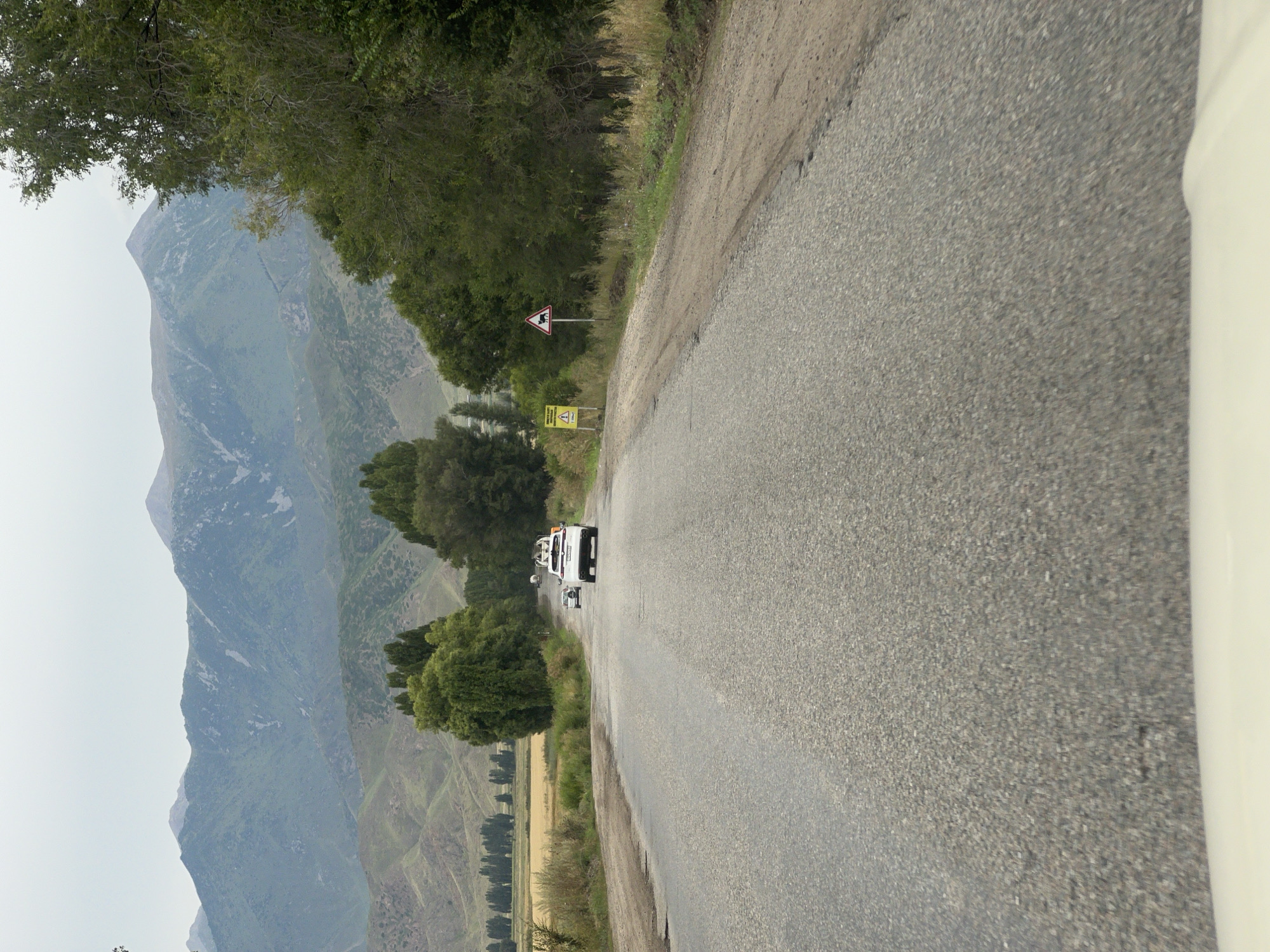 The image depicts a road in Balbay, Kyrgyzstan, leading towards a mountainous landscape. The road is paved and shows signs of wear and tear. On the right side of the road, there is a dirt shoulder with dry grass and vegetation. Further down the road, two vehicles are visible: a white SUV with luggage on its roof and another white car. Trees line both sides of the road, with a field of golden crops in the distance on the left, bordered by a line of tall trees. The background is dominated by a series of rolling green hills, with more rugged, bare mountains in the far distance, their peaks partially obscured by a cloudy sky. Two yellow warning signs are present on the right side of the road, one with a symbol resembling a person and another with a symbol of livestock.