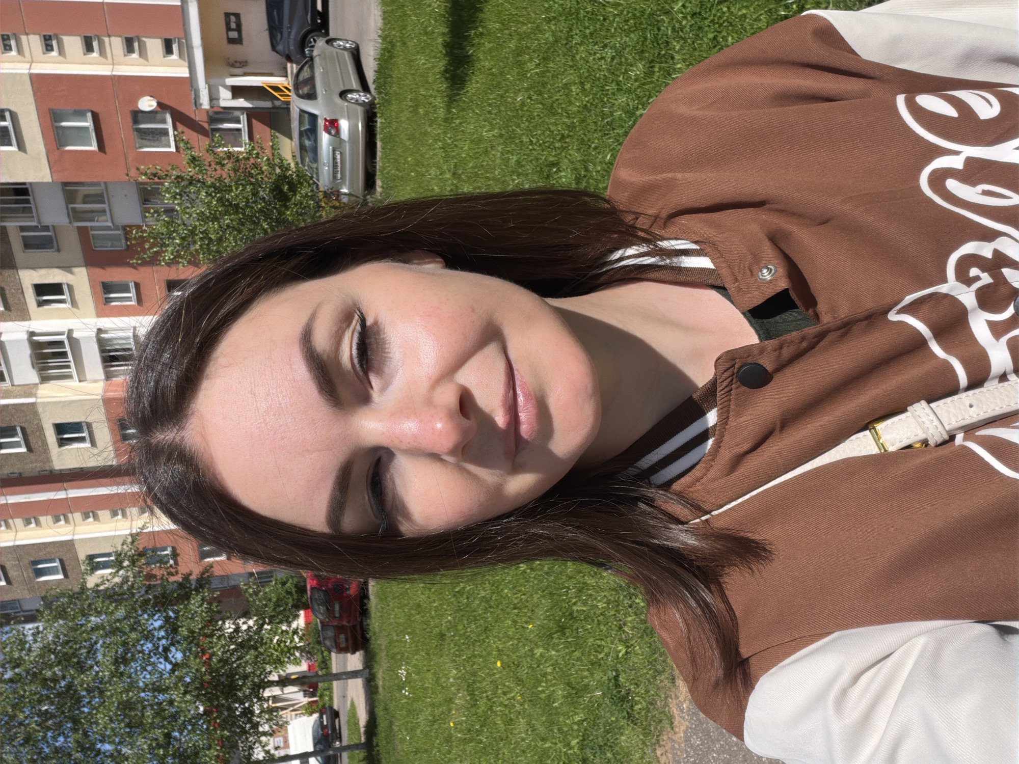 A young woman poses outdoors in the city of Unknown, Unknown. She wears a brown and white varsity jacket, with a light-colored strap across her shoulder. Her hair is brown. The background includes a building with multiple windows and a green lawn. Cars are parked nearby, and trees are visible. The lighting suggests a sunny day.