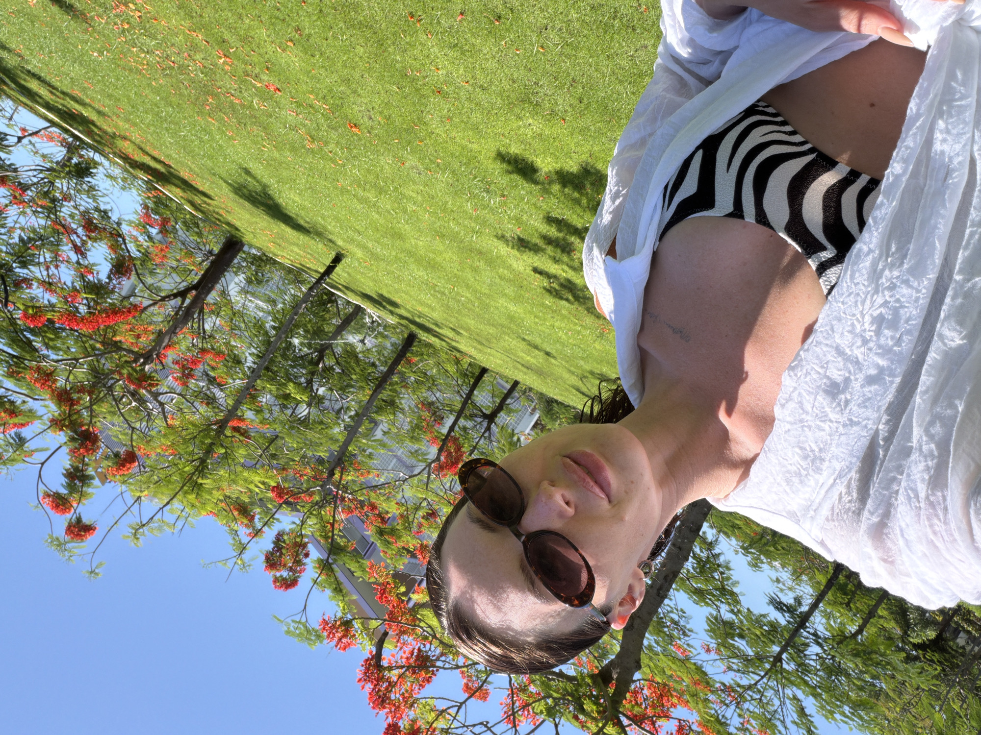 A woman is captured in a close-up, slightly tilted shot outdoors, with a bright blue sky visible behind her. She is wearing sunglasses with tortoise-shell frames and a black and white zebra-print bikini bottom, partially covered by a loose white garment. Her skin appears to have some light markings or tattoos. The background features lush green grass and the branches of a tree adorned with vibrant red flowers, suggesting a tropical or subtropical environment. Sunlight casts shadows on the grass, indicating it is likely daytime. The overall mood appears relaxed and casual, with the woman seemingly enjoying a sunny day in a natural setting. The specific location is Dong Ninh Hoa Ward, Vietnam.