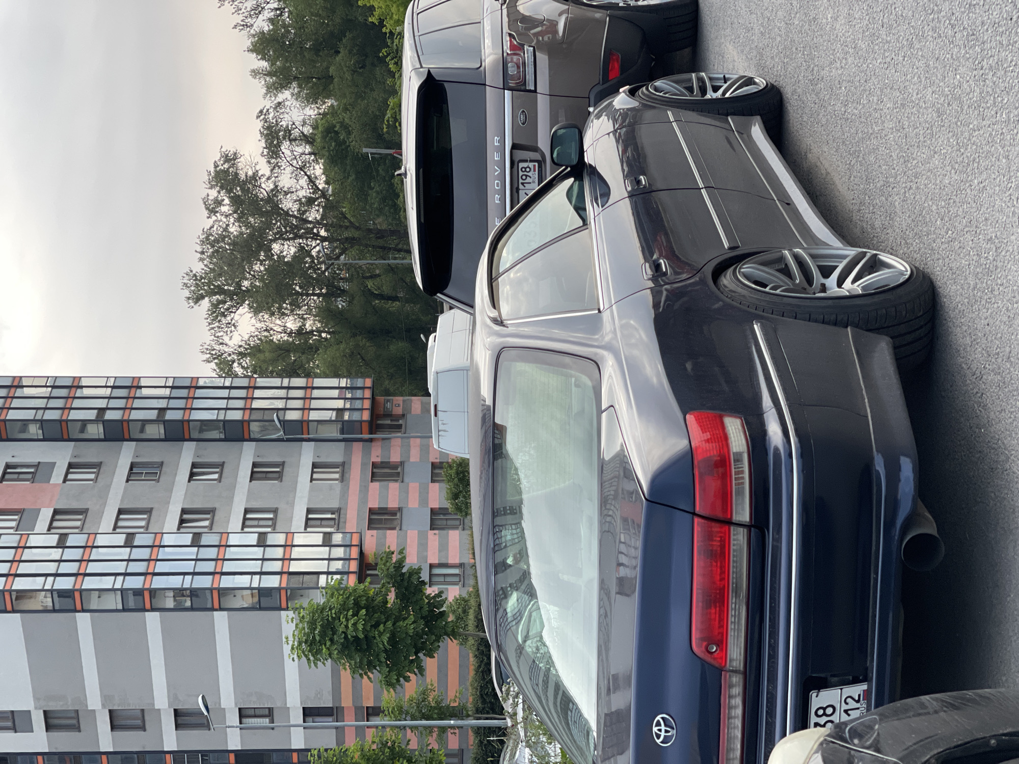 A dark blue sedan, possibly a Toyota Mark II, is parked in the foreground, facing left. The car has a clean exterior and aftermarket wheels. To its right, a brown Range Rover is parked, also facing left. The Range Rover's license plate reads "198 RUS". Behind the cars, a modern, multi-story building with a gray and orange facade stands prominently. Trees and other vegetation are visible around the building and behind the vehicles. The sky is overcast. The image appears to be taken outdoors during the daytime.