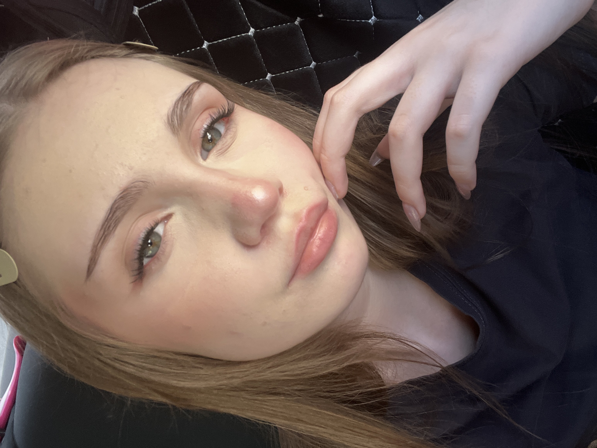 The image is a close-up, head-and-shoulders selfie of a young Caucasian female with light brown hair. She is wearing a dark-colored, V-neck shirt. Her makeup includes blush on her cheeks and nose, eyeliner, and lip gloss. She has her right hand resting on her chin and cheek, with her fingers slightly curled. Her fingernails are painted a light, neutral color. The background appears to be a dark, quilted surface on the right and a dark, blurred area on the left, possibly an interior setting. There are no discernible landmarks indicating a specific location within Sterlitamak, Russia.