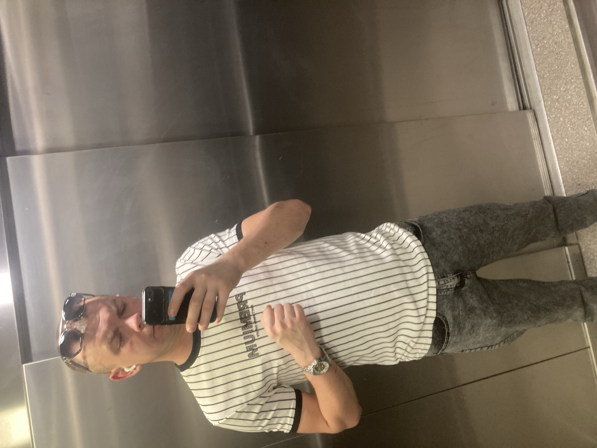 A man is taking a selfie in an elevator. He is wearing sunglasses on his head, a white and black striped t-shirt, gray jeans, and a wristwatch. He is holding a black smartphone. The interior of the elevator is made of stainless steel. There is no evidence to suggest the location is Khimki, Russia.