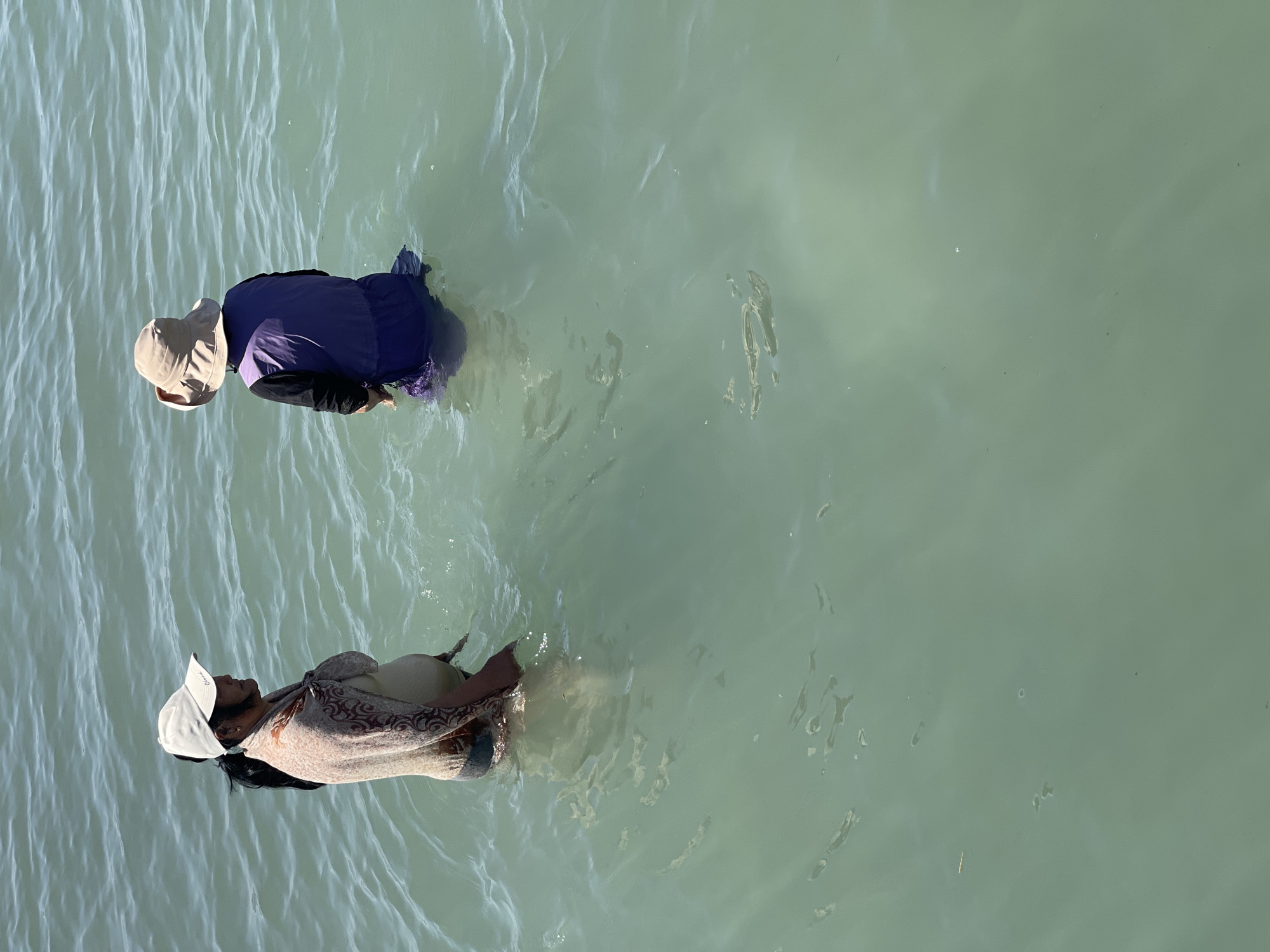 Two individuals are standing in shallow, greenish water, likely a lake or a slow-moving river. The person on the left is facing right in profile, wearing a cream-colored baseball cap, a patterned wrap or shawl over a light-colored top, and dark bottoms. Their hair is dark and pulled back. The person on the right is facing away from the camera, wearing a wide-brimmed beige hat and a purple vest over a long-sleeved dark garment and dark bottoms. The water has a rippled surface, indicating gentle movement. The scene appears to be outdoors during daylight, with the water reflecting the sky. The image does not provide any specific visual cues that would allow for pinpointing the location within Ornek, Kyrgyzstan.