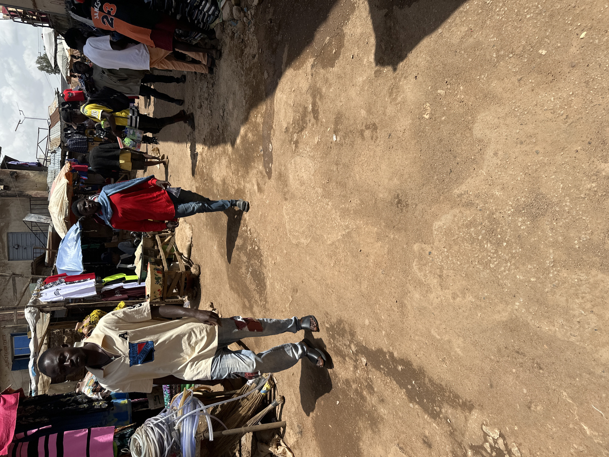 The image depicts a bustling market scene in Bukuru, Nigeria. In the foreground, a man wearing a tan polo shirt and light blue jeans walks towards the camera, his feet clad in black sandals. To his left, a stall displays various items, including coiled hoses and bundles of broomsticks. Further into the market, other individuals are visible browsing stalls and moving through the dirt-covered pathway. Stalls with corrugated metal roofs line the sides of the path, displaying clothing and other goods. The environment appears to be a typical open-air market with a mix of commerce and pedestrian traffic under a partly cloudy sky.