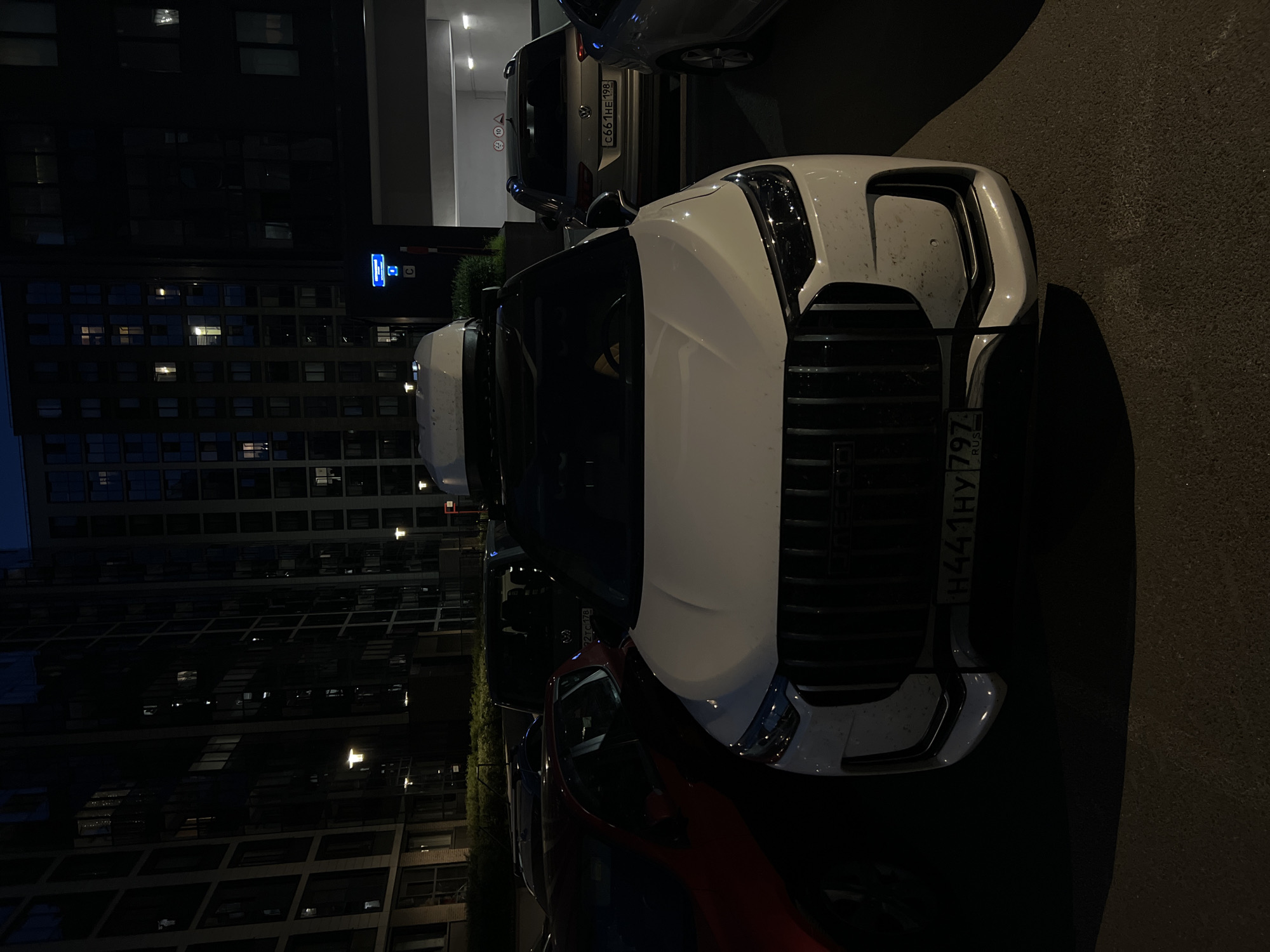 The image captures a night scene in a city, likely a residential or mixed-use area in Russia, given the visible license plates. The primary focus is a white SUV parked in what appears to be an outdoor parking lot. The SUV has a prominent front grille and a roof-mounted cargo box. It is flanked by other vehicles, including a red car to its left and a silver SUV to its right. In the background, tall apartment buildings with illuminated windows are visible, suggesting the presence of a dense urban environment. The lighting is dim, with streetlights and the building lights providing the illumination. The overall atmosphere is dark and somewhat indistinct, typical of a nighttime urban setting. The license plate of the white SUV reads "H441HY 797 RUS". The license plate of the silver SUV reads "C661HE 198". There are no people visible in the frame.