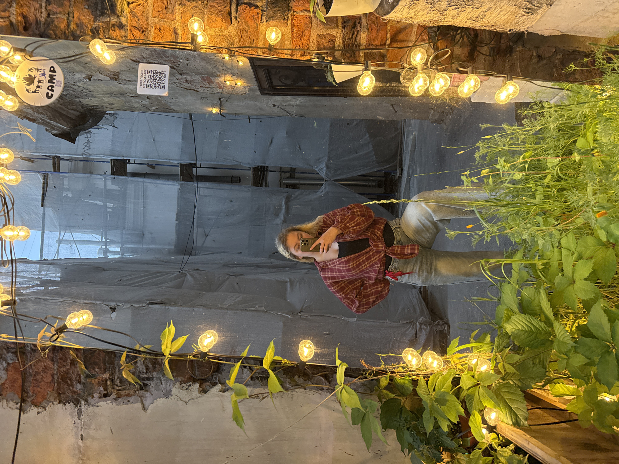 A young woman with blonde hair is taking a mirror selfie in an alleyway. She is wearing a red and white plaid shirt over a black top and light-colored jeans. String lights with Edison bulbs are draped along the brick walls and over some greenery, creating a warm and inviting atmosphere. The alley appears to be under construction or renovation, as indicated by the grey tarpaulin covering some structures. There is a round sticker on the wall with the word "CAMP" on it and a QR code. The overall mood is casual and perhaps a bit adventurous, given the urban setting and the string lights. The image seems to be taken in the evening or at night, as the lights are prominently illuminated. The location is Saint Petersburg, Russia.