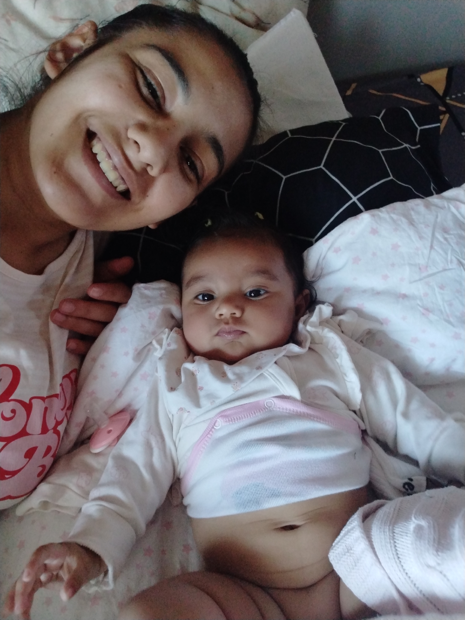 A woman and a baby are lying on a bed with patterned pillows and a white blanket. The woman is smiling at the camera. The baby is lying on its back, looking at the camera. The baby is wearing a white outfit with a pink trim. The location is Unknown, Unknown.