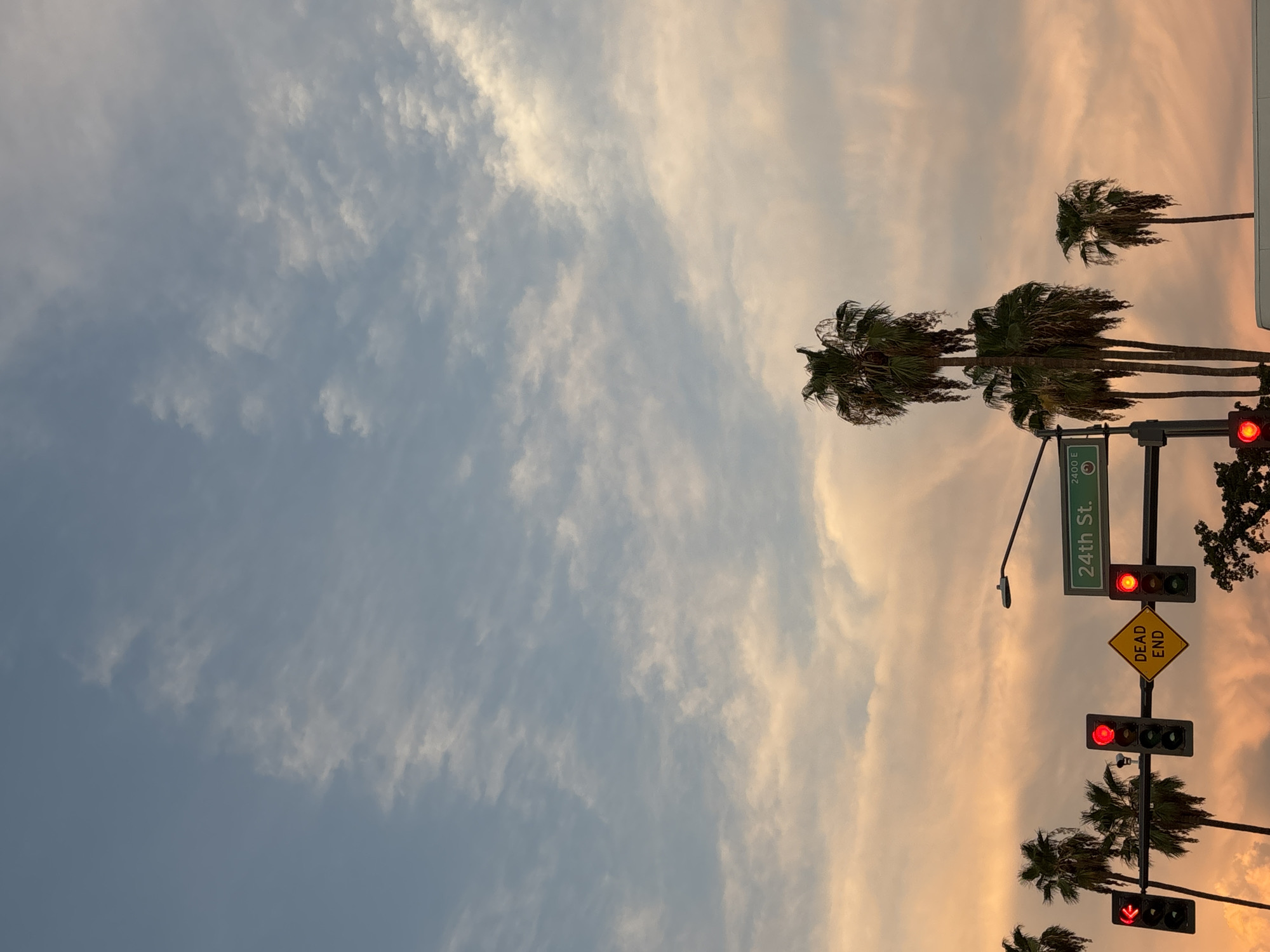 The image shows a street scene in Phoenix, Arizona, USA, at sunset. Palm trees are silhouetted against a sky with soft pink and orange clouds. Traffic lights, a street sign indicating "24th St.", and a "Dead End" sign are visible. The overall atmosphere is calm and serene, with the warm colors of the sunset creating a picturesque scene.