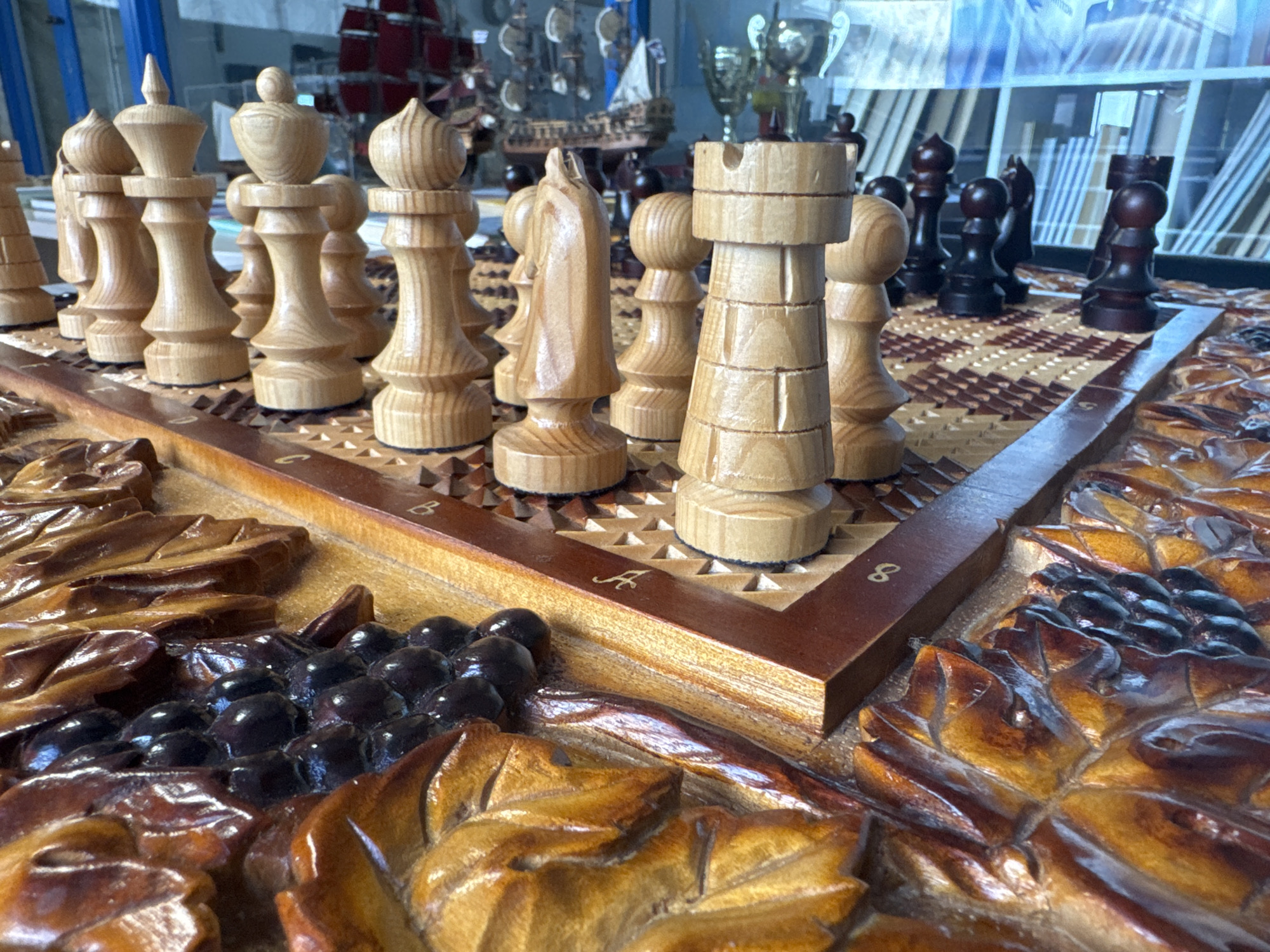 The image shows a close-up of a carved wooden chess set. The board features detailed carvings of grapes and leaves around the playing surface. The chess pieces are made of light-colored wood and are arranged on the board. In the background, a window reflects a cityscape and a model sailboat and some trophies can be seen on display. According to the source image's metadata, it was taken in Saint Petersburg, Russia.