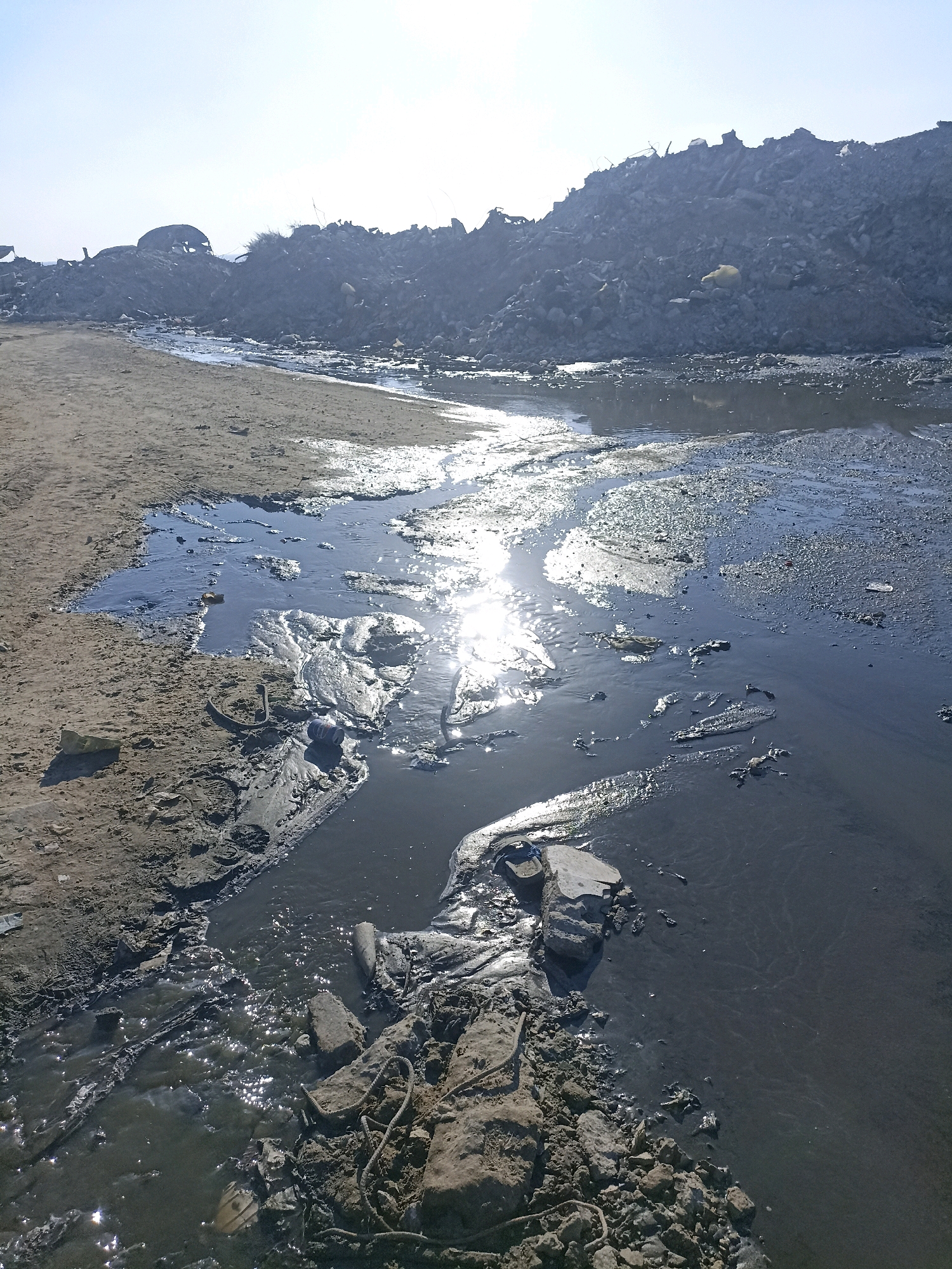 This image depicts a polluted waterway in the city of Gaza, Palestinian Territories. A shallow stream of dark, possibly sewage-tainted water flows through a sandy and rocky embankment. The water reflects the bright sunlight, creating shimmering highlights. Large piles of debris and rubble dominate the background, suggesting a heavily impacted or industrialized area. Some plastic waste is visible on the sandy bank. The sky is clear and light blue.