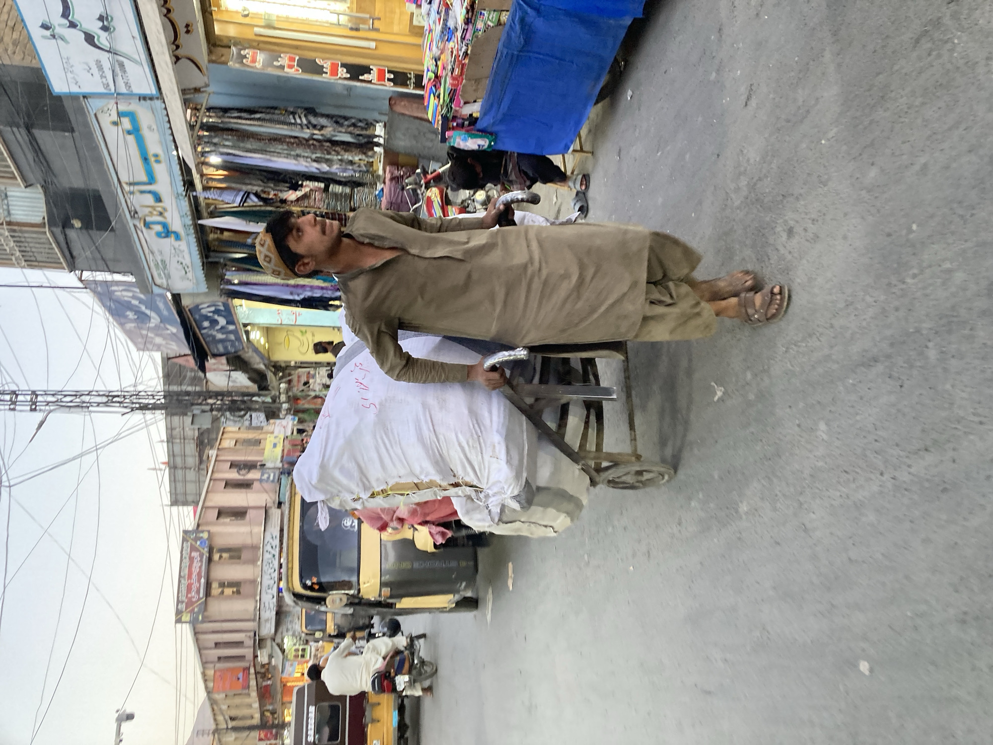 A man wearing a light brown shalwar kameez and a brown and tan patterned cap is pulling a handcart loaded with white and multi-colored packages. He is wearing sandals. The background shows a street scene with various shops and buildings. The scene is taken during daytime. The image likely depicts a scene in Quetta City Tehsil, Pakistan.