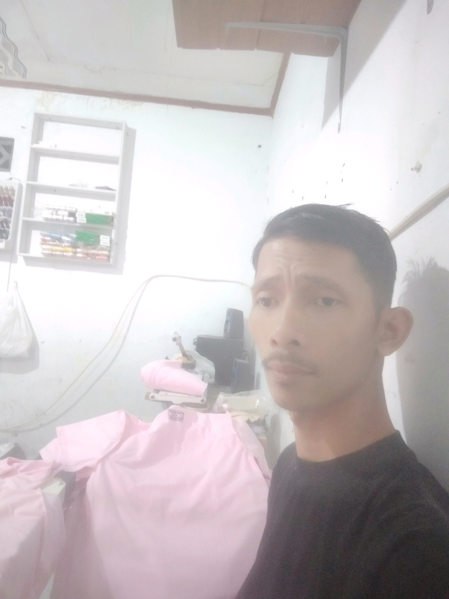 A man is taking a selfie in what appears to be a workshop or tailor shop in Cikoneng, Indonesia. The man is wearing a dark t-shirt and has a mustache. In the background, there is a shelf with spools of thread, a sewing machine, and pink fabric. The walls are white and there is a wooden ceiling.