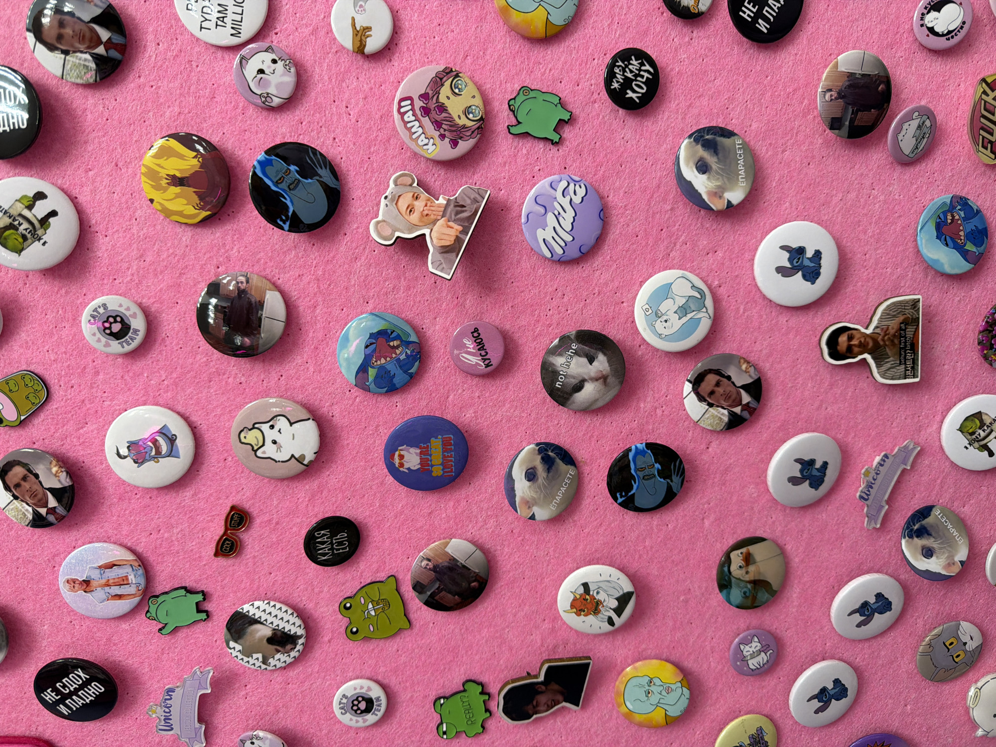 The image shows a collection of approximately 60 small, mostly round badges and pins scattered on a pink felt surface. The badges feature a variety of images and text, including:
* Illustrations of cartoon characters, such as Stitch from Lilo & Stitch, characters from Hercules (Hades), and various animals like cats, dogs, and frogs.
* Images of people, including celebrities and possibly personal photos.
* Text in both English and Russian, with phrases like "KAWAII," "FUCK," "Unicorn," and "Я не Кусаюсь" (I don't bite).
* Various objects such as sunglasses.
The badges vary in size and shape, with most being round but some being custom-shaped to match the image they depict. They are arranged in a seemingly random pattern across the pink felt surface. The pink felt shows some subtle texture.