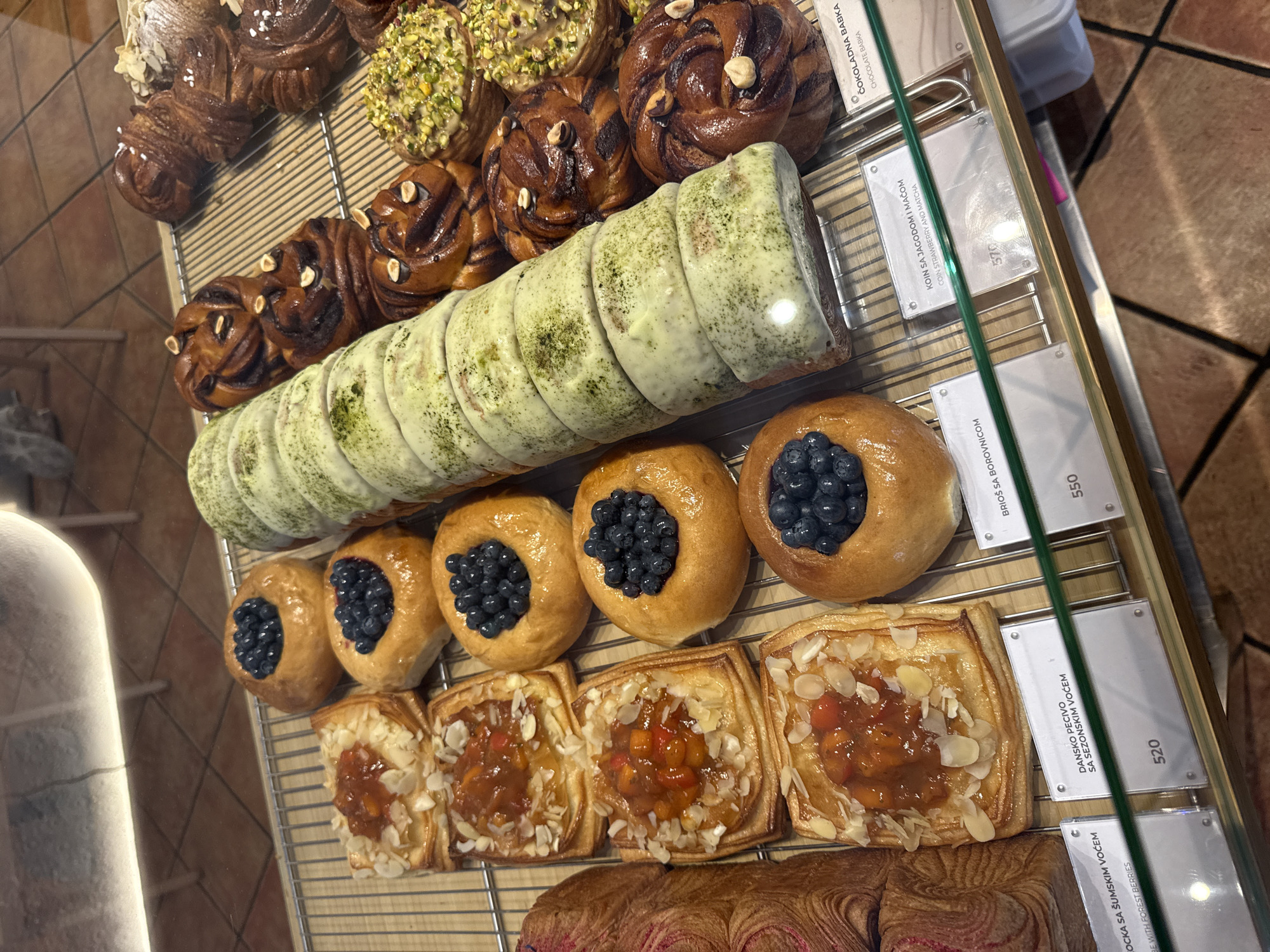 The image shows a display case full of various pastries in a bakery. The pastries include fruit tarts topped with almonds, buns filled with blueberries, and cinnamon rolls. There are also some larger, cylindrical pastries with a green-colored coating, possibly matcha flavored. The pastries are arranged on metal racks. Labels with names and prices in a foreign language are visible in front of some of the items. The background shows a tiled floor and a white, illuminated counter. The setting appears to be a modern bakery, possibly in Belgrade, Serbia, given the Cyrillic script on some of the labels. There are no people visible in the image.