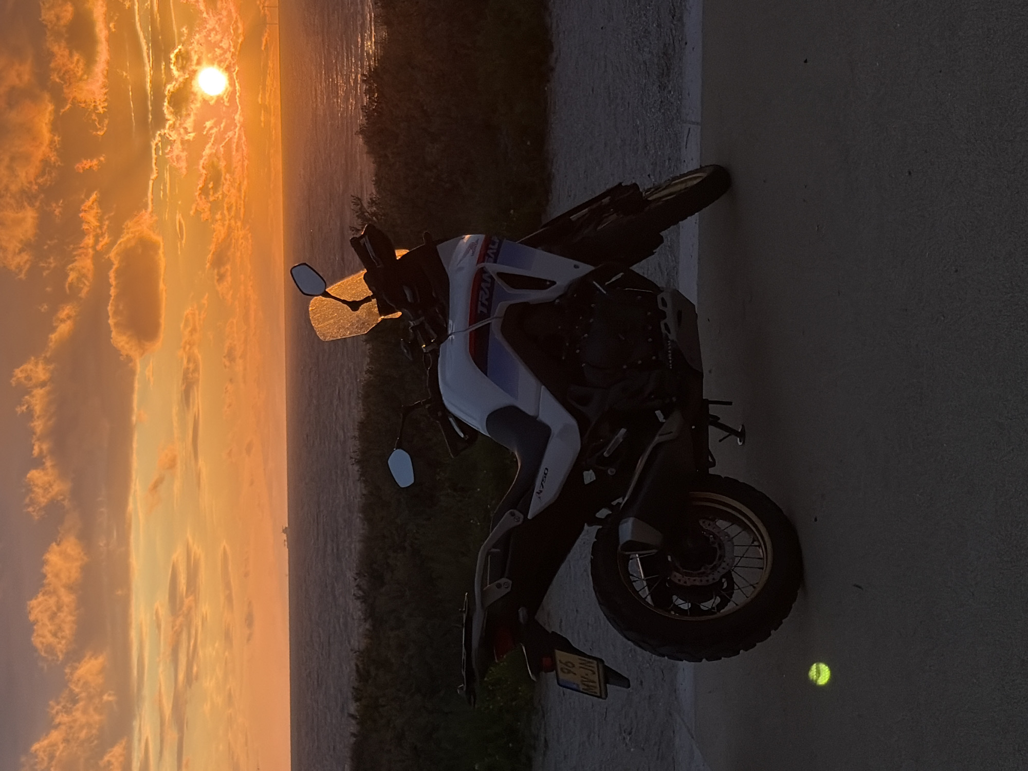 A motorcycle is parked on a paved surface near the coast of The Hague, Netherlands, at sunset. The sky is filled with vibrant orange and yellow clouds, with the sun partially obscured by clouds and reflecting on the water. The motorcycle, a white Honda Transalp 750, is positioned facing left, with its license plate visible. A small, bright yellow object, possibly a tennis ball, is on the ground in front of the motorcycle. In the background, the ocean stretches to the horizon, with a distant ship and some wind turbines visible. The scene is dimly lit by the setting sun, casting shadows and creating a silhouette effect on the motorcycle and surrounding vegetation.
