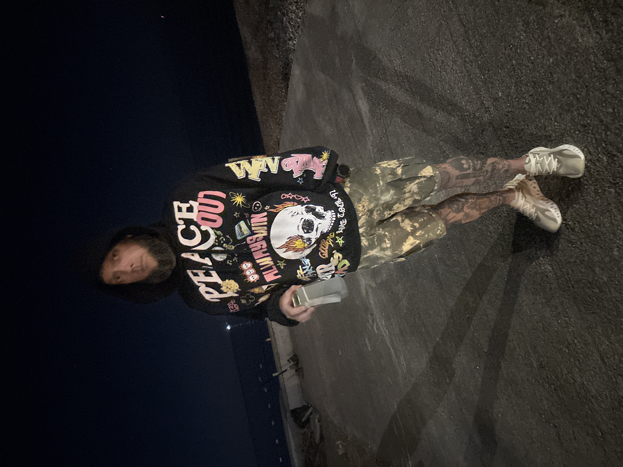 A person is standing on pavement with dark asphalt in the city of Bar, Montenegro. The person wears a black hoodie with graphics, camouflage shorts, and light gray shoes. They are holding a small, rectangular object in their hand. The background shows a dark night sky and the corner of a wall or building. The image seems to be taken from a low angle. The person's legs and arms are tattooed.