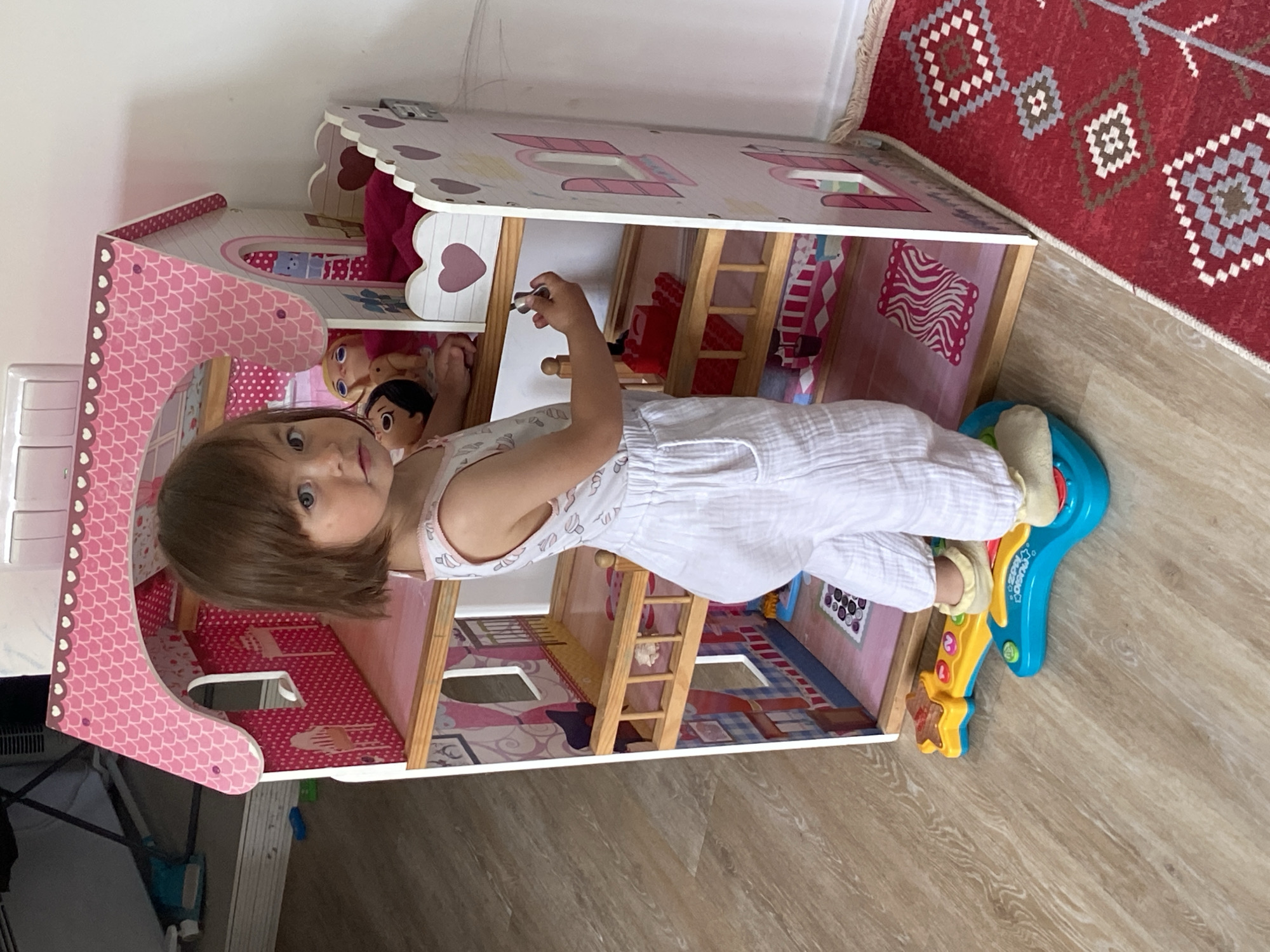 A young girl in white pajamas is playing with a pink dollhouse filled with toys in a room. The room has light wood floors and is adjacent to a red rug with geometric patterns. A toy guitar is on the floor near the dollhouse. The location is Tyumen, Russia.