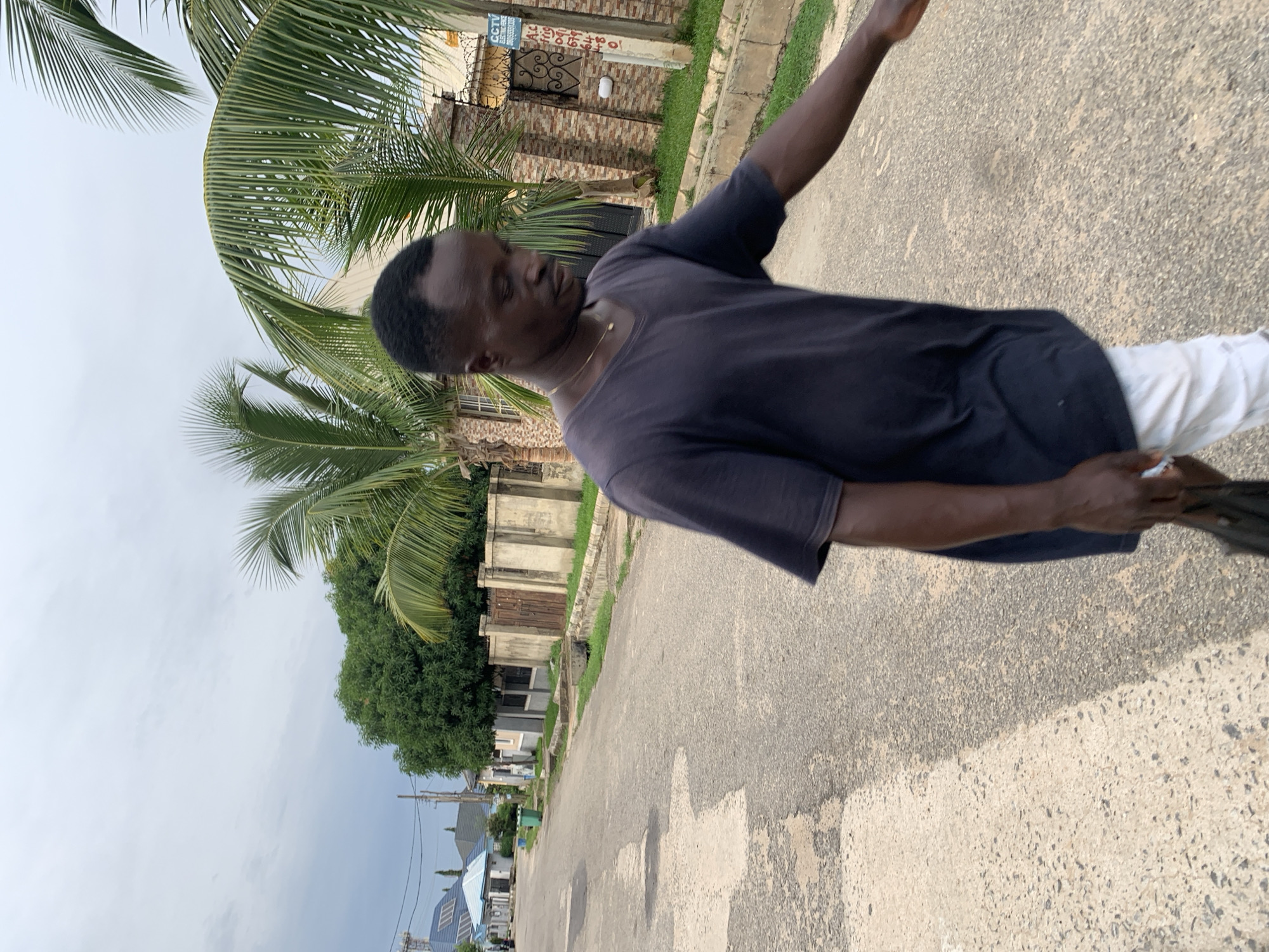 An African man wearing a dark v-neck t-shirt and light-colored pants walks on a paved road in Abuja, Nigeria. He is carrying a dark bag in his left hand. Palm trees and residential buildings line the street. The sky is overcast.
