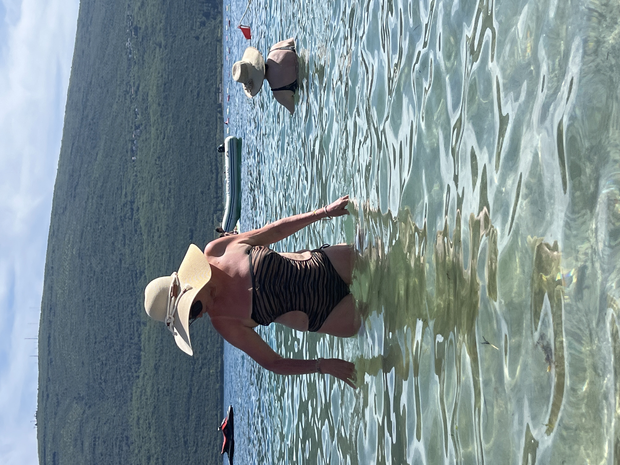 The image depicts a sunny day at a beach in Baošići, Montenegro. In the foreground, a woman wearing a wide-brimmed straw hat and a black and brown tiger-striped one-piece swimsuit stands in clear, shallow water. She has her right arm extended, hand slightly submerged, and her left arm is bent. Behind her, another woman, also wearing a straw hat, faces away from the camera, partially submerged in the water. In the background, a verdant, tree-covered mountain slopes down towards the water, with a few scattered buildings visible on its side. Further out in the water, a small inflatable boat with the text "TENDER TO LANA" is present, as well as a red buoy. A jet ski is also visible on the left side of the image. The water is calm with gentle ripples, and the sunlight creates reflections on the surface.