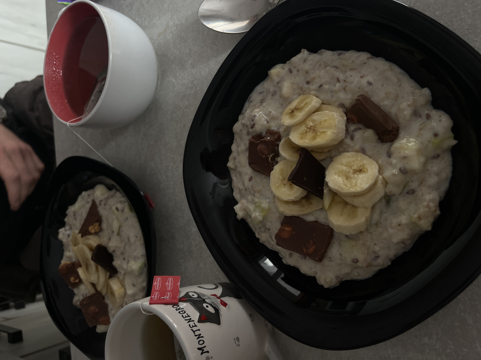 The image shows two bowls of porridge topped with banana slices and chocolate chunks, along with two mugs of tea. The scene is set on a grey surface. A spoon is visible near the larger bowl. One of the mugs has a black cat design and the word "MONTENEGRO" on it, alongside a teabag label indicating the location is Bečići, Montenegro. A person's arm is partially visible on the left side of the image.