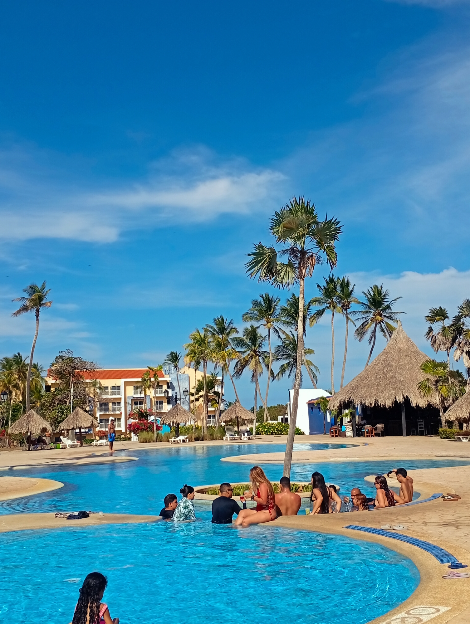 The image depicts a resort swimming pool in Playa El Agua, Venezuela. A group of approximately 15 people are either in the pool or sitting along its edge. Some are interacting with each other, while others are looking out toward the pool or the surrounding area. The pool is surrounded by palm trees and thatched-roof structures, some of which appear to be bars or relaxation areas. In the background, there is a multi-story building with balconies. The sky is blue with scattered clouds, indicating a sunny day.