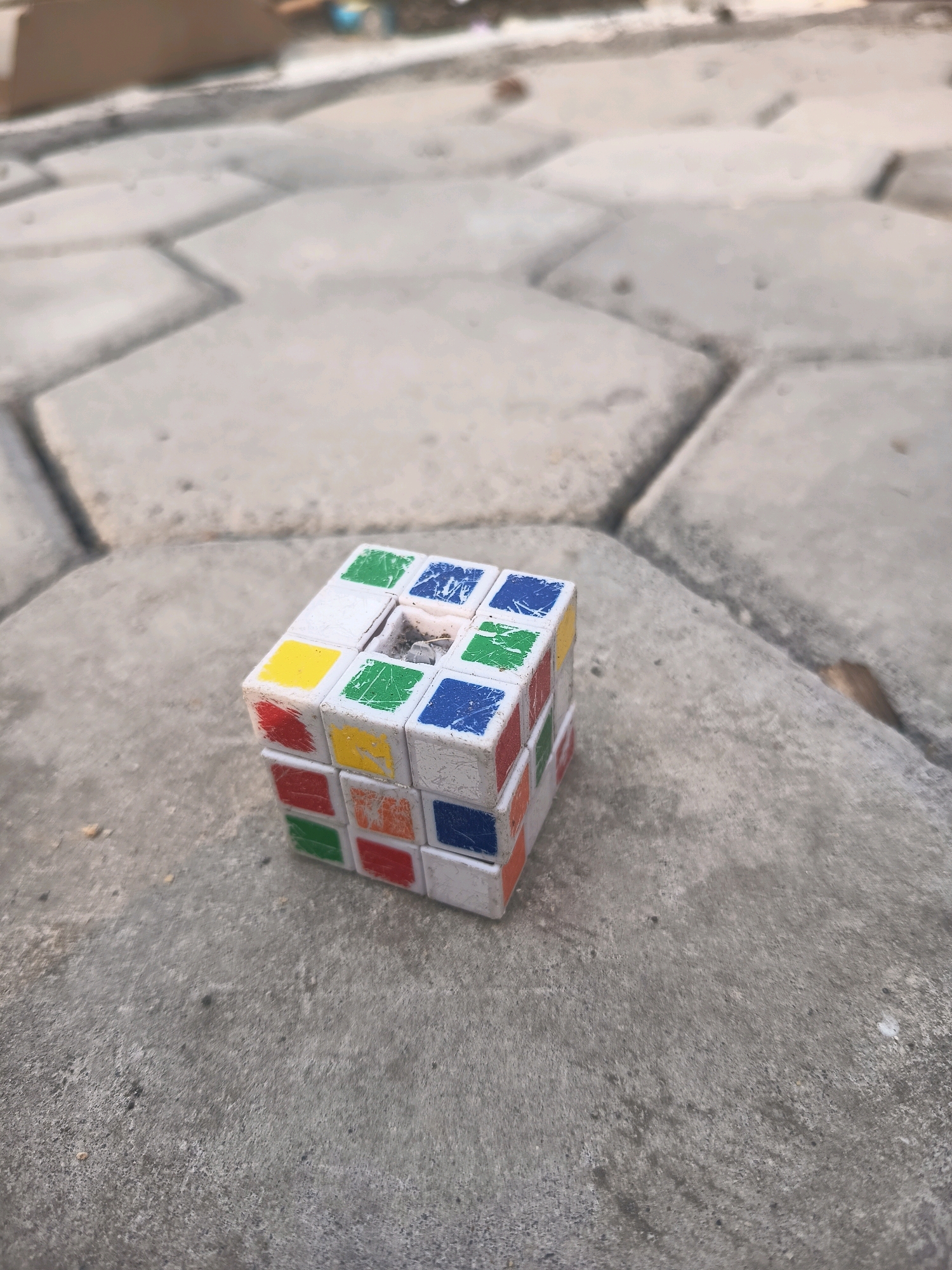 A close-up, medium-angle, outdoor shot shows a damaged Rubik's Cube lying on a light gray, textured concrete surface. The concrete is comprised of large, irregularly shaped interlocking pavers with visible grout lines. The Rubik's Cube, a standard 3x3 puzzle, is missing several colored squares and shows signs of wear, with scratches and chipped paint on its white plastic casing. The visible colors on the cube are red, orange, yellow, green, and blue. In the background, out of focus, is a glimpse of what appears to be construction materials or debris, and a small portion of a blue object. The lighting suggests it is daytime. The location is identified as Sidorejo, Indonesia.