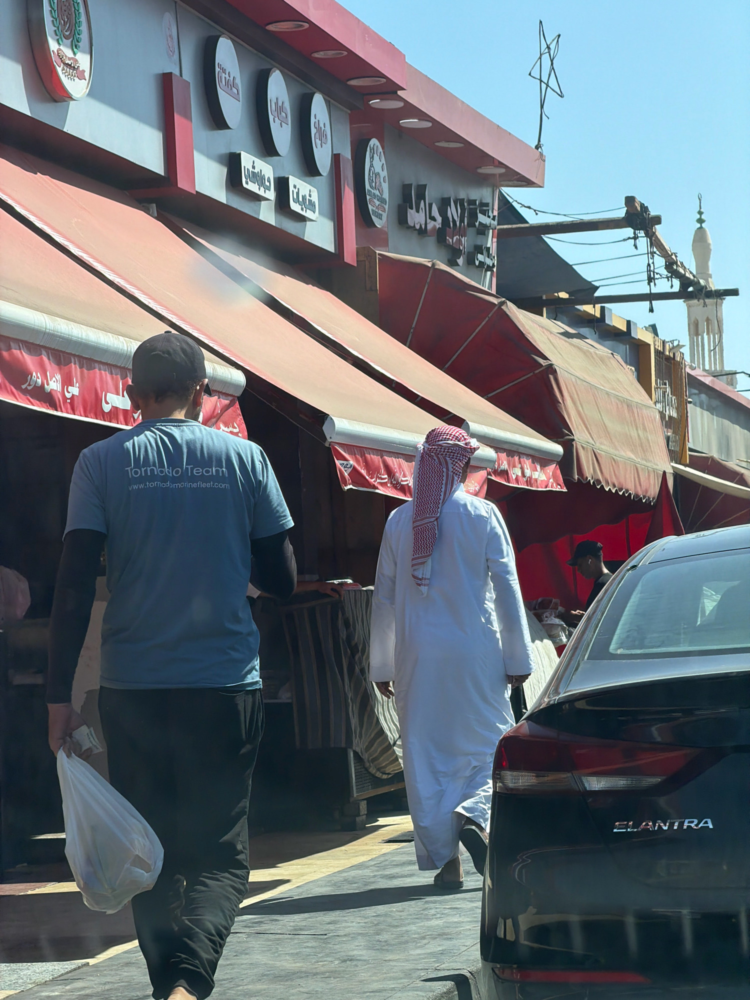 In an outdoor urban setting, likely Sharm El Sheikh, Egypt, two men are walking away from the camera on a paved surface. The man on the left wears a light blue t-shirt with the text "Tornado Team" and "www.tornadomarinefleet.com" printed on the back, black long sleeves, and black pants. He carries a white plastic bag filled with items in his right hand. The man on the right is dressed in a traditional white thobe and a red and white checkered ghutra. They are walking past a building with red awnings and Arabic signage. A black Hyundai Elantra car is partially visible on the right side of the frame, with the "ELANTRA" badge clearly displayed on its trunk. In the background, a minaret of a mosque is visible against a clear blue sky. The scene appears to be a street or sidewalk in front of a commercial establishment.