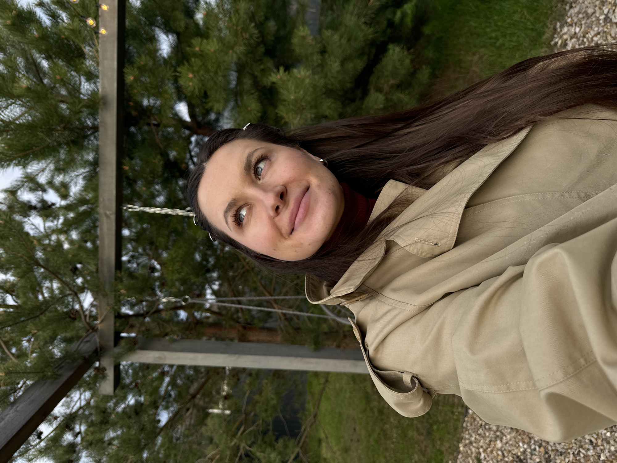 A woman with long brown hair and green eyes is posing for a selfie. She is wearing a beige trench coat over a red turtleneck sweater. Her hair is partially obscuring the right side of her face and shoulder. She is standing next to a swing set in an outdoor area with evergreen trees in the background. The ground appears to be covered in gravel. The location is Литвиново, Russia.