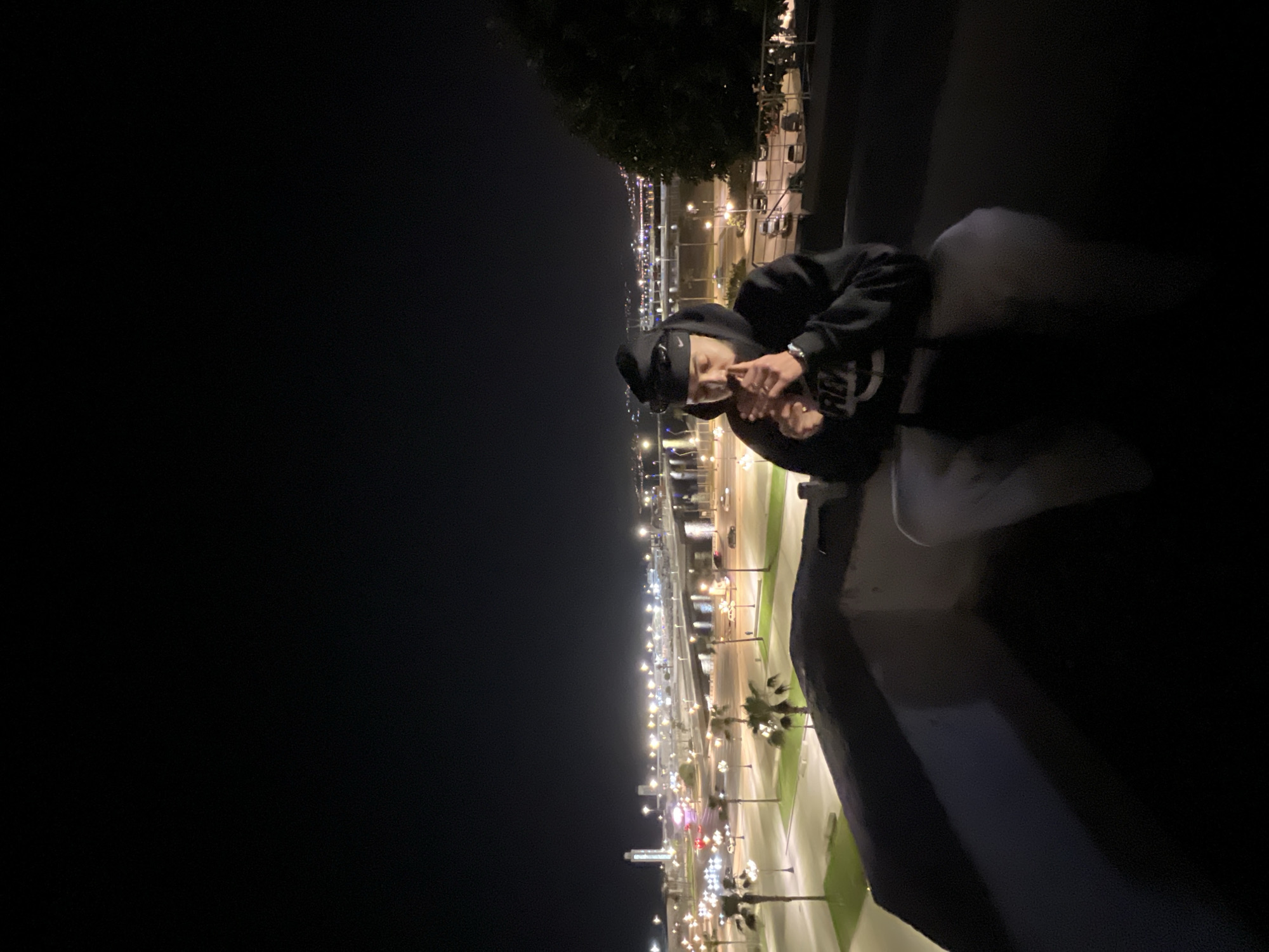 The scene depicts a male individual at night, viewed from an elevated position overlooking the city of Tanger, Morocco. The man is situated on a dark, low parapet or wall in the foreground. He is wearing a black hooded sweatshirt featuring a light-colored graphic on the chest, light-toned pants, a dark baseball cap with a Nike logo, and sunglasses resting on the cap. A wristwatch is visible on his left wrist. His hands are raised to his face, actively engaged in lighting a small, cylindrical object, consistent with a cigarette. The background displays an extensive, brightly illuminated urban and port landscape. This area includes a multi-lane roadway network, numerous streetlights, and vehicle lights (headlights and taillights) indicating active traffic. Palm trees are visible along the illuminated streets. On the far left of the background, a prominent white, cylindrical tower with a rounded top, consistent with a port control tower, rises above the port area. The illuminated city extends across a visible body of water into the distance.