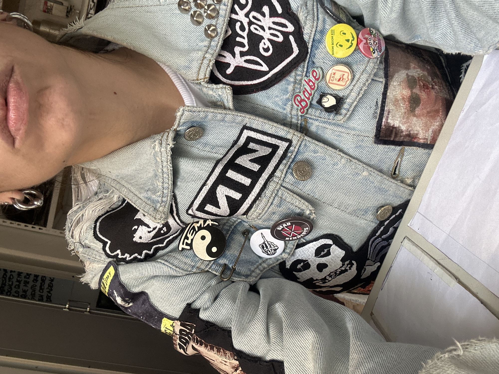 The image shows a person wearing a light blue denim jacket adorned with various patches and buttons. The patches include text like "fuck off," "NIN," and symbols such as a yin-yang. Buttons feature a smiley face and other designs. The person's face is partially visible, showing lips, neck, and an earring. Behind the person, a shelf with assorted items is visible, including a poster or sign with printed text. The image is taken indoors with natural light. The city of Buenos Aires, Argentina is not discernible from the details provided.