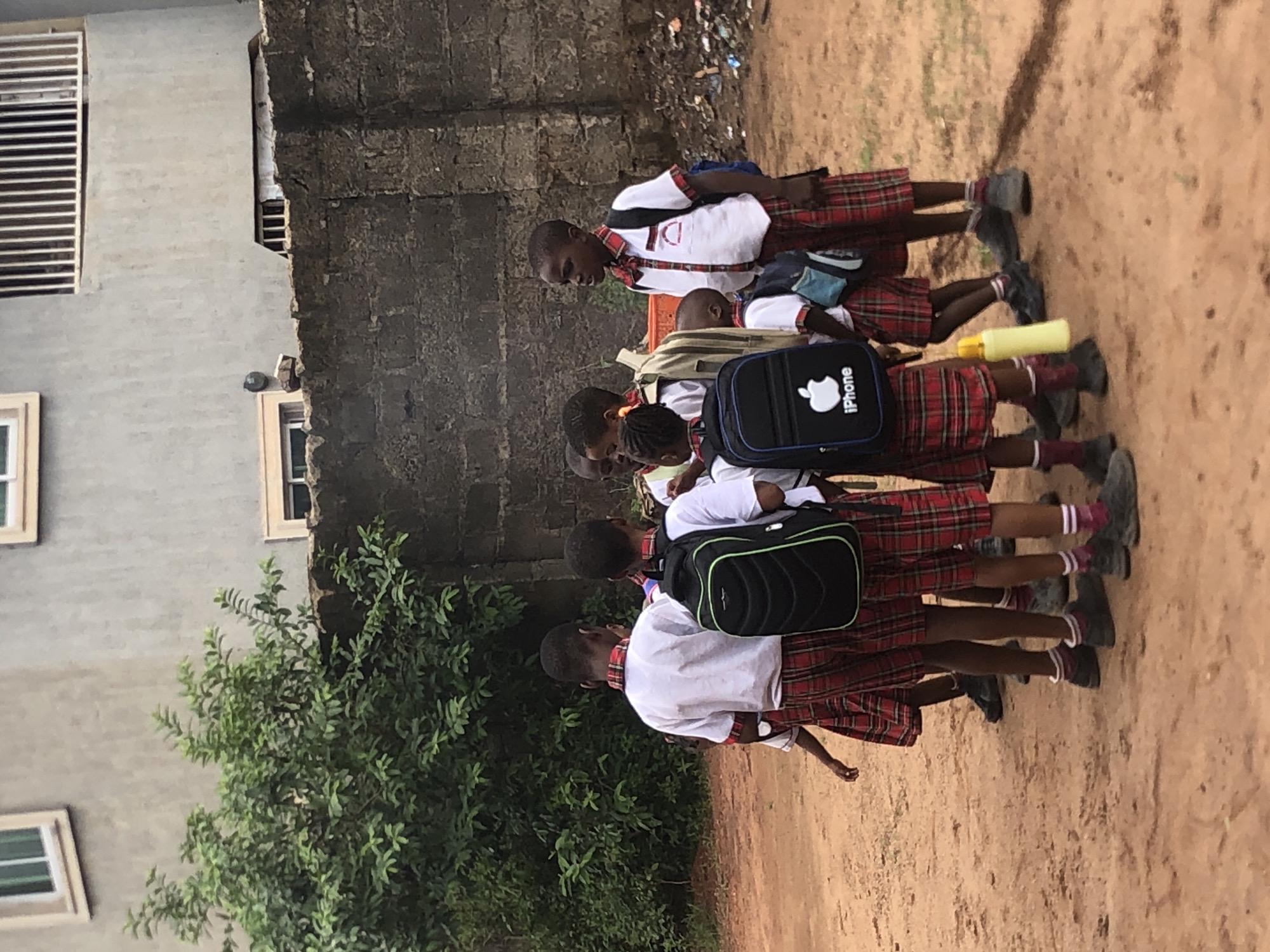 The image displays a group of young school children, mostly seen from behind, walking in a line on a dirt path in Ukwu-Abwa, Nigeria. They are uniformly dressed in white short-sleeved shirts, red and black plaid skirts or shorts, and dark shoes with white and red/maroon striped socks.
The children are carrying various school bags. One prominent black backpack features a white Apple logo and the word "iPhone" printed beneath it. Another child has a black backpack with green accents. A yellow water bottle is visible, held by one of the children.
In the background, to the left, is a light-colored building with several windows, some featuring horizontal louvered vents. To the right, a rough, dark-textured wall stands, with what appears to be scattered debris or refuse beyond it. A vibrant green bush or small tree is positioned between the path and the buildings. The scene appears to take place during the daytime under natural light. The children are engaged in the activity of walking, possibly during a school commute.