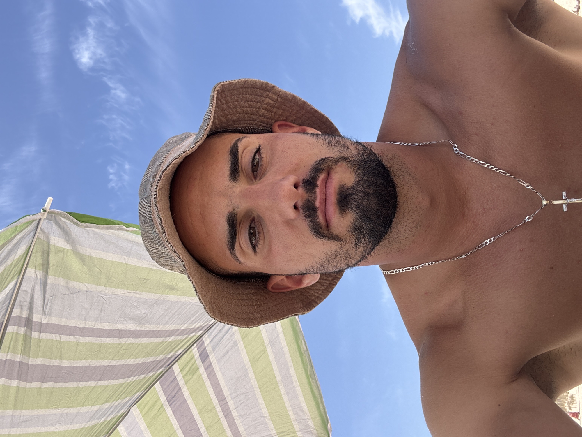 A low-angle shot looking up at a man wearing a brown bucket hat and silver chain with a cross pendant. The man has a dark beard and is shirtless. The background shows a blue sky with wispy clouds and a striped beach umbrella in shades of green and white. The location is Unknown, Unknown.