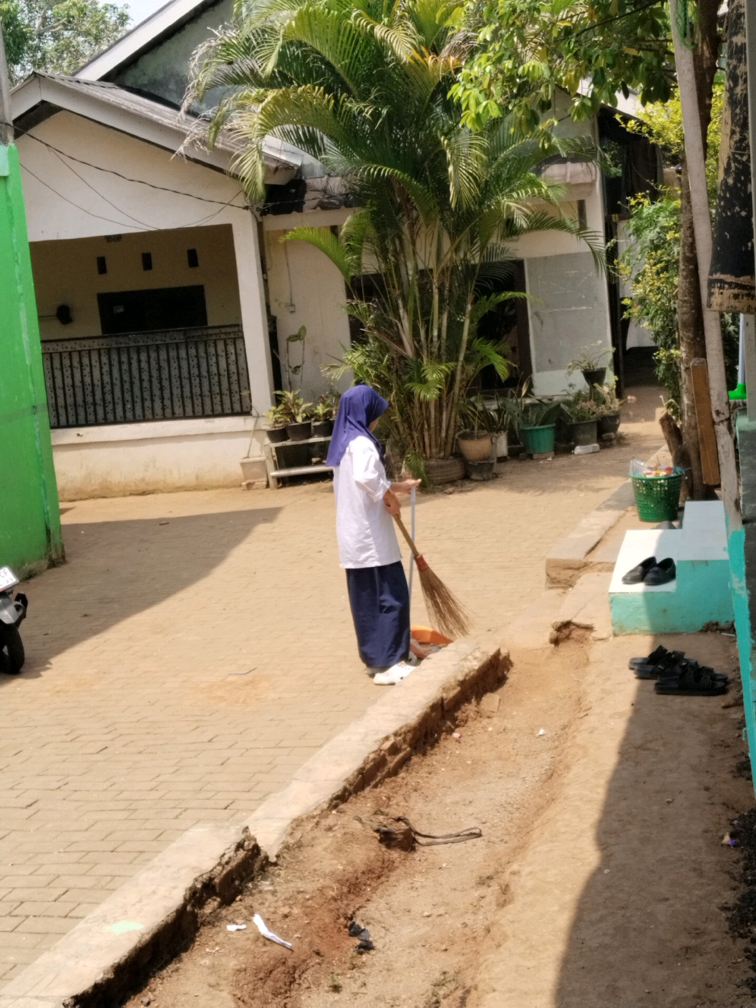 A young woman wearing a blue hijab, white shirt, and blue pants sweeps a sandy courtyard with a broom. She is standing next to a shallow, dirt-filled trench with a concrete edge. Behind her, a white house with a dark-colored metal fence and a large palm tree are visible. To her right, there is a green trash can, a light blue step, and several pairs of black sandals. A motorcycle is parked to the left of the frame. The scene appears to be in a residential area with buildings and vegetation under a bright, sunny sky.