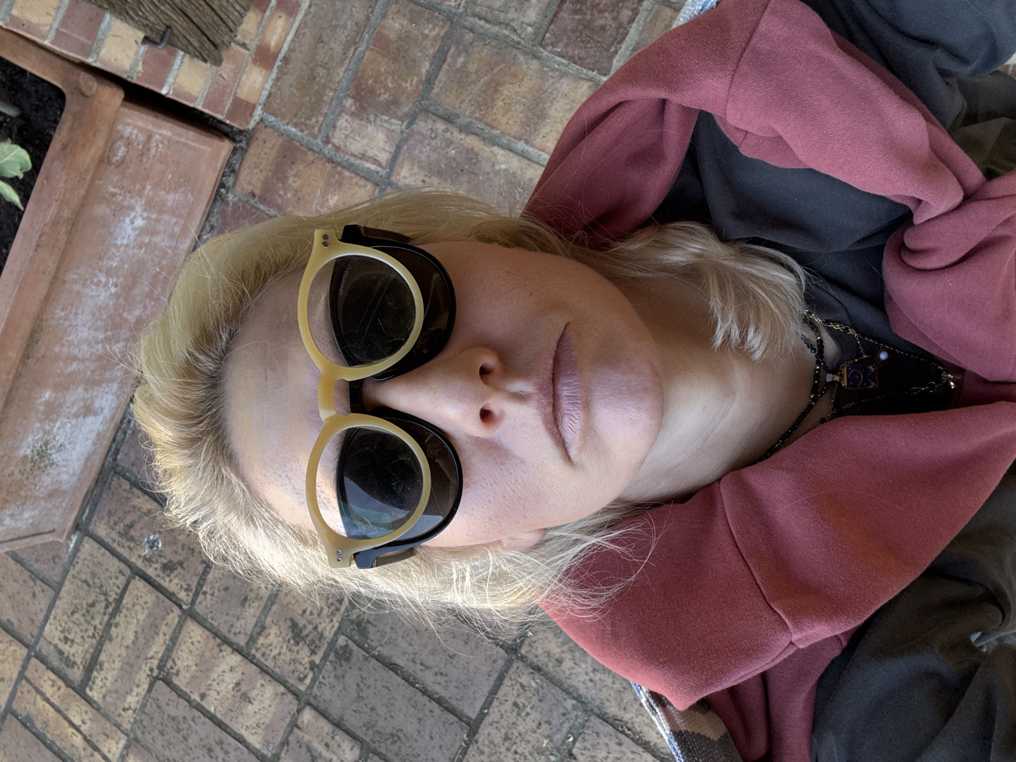 A woman with blonde hair wearing sunglasses and a maroon hoodie is lying on a brick surface, possibly a patio or walkway. A potted plant is visible in the upper left corner. The location is in Velletri, Italy.