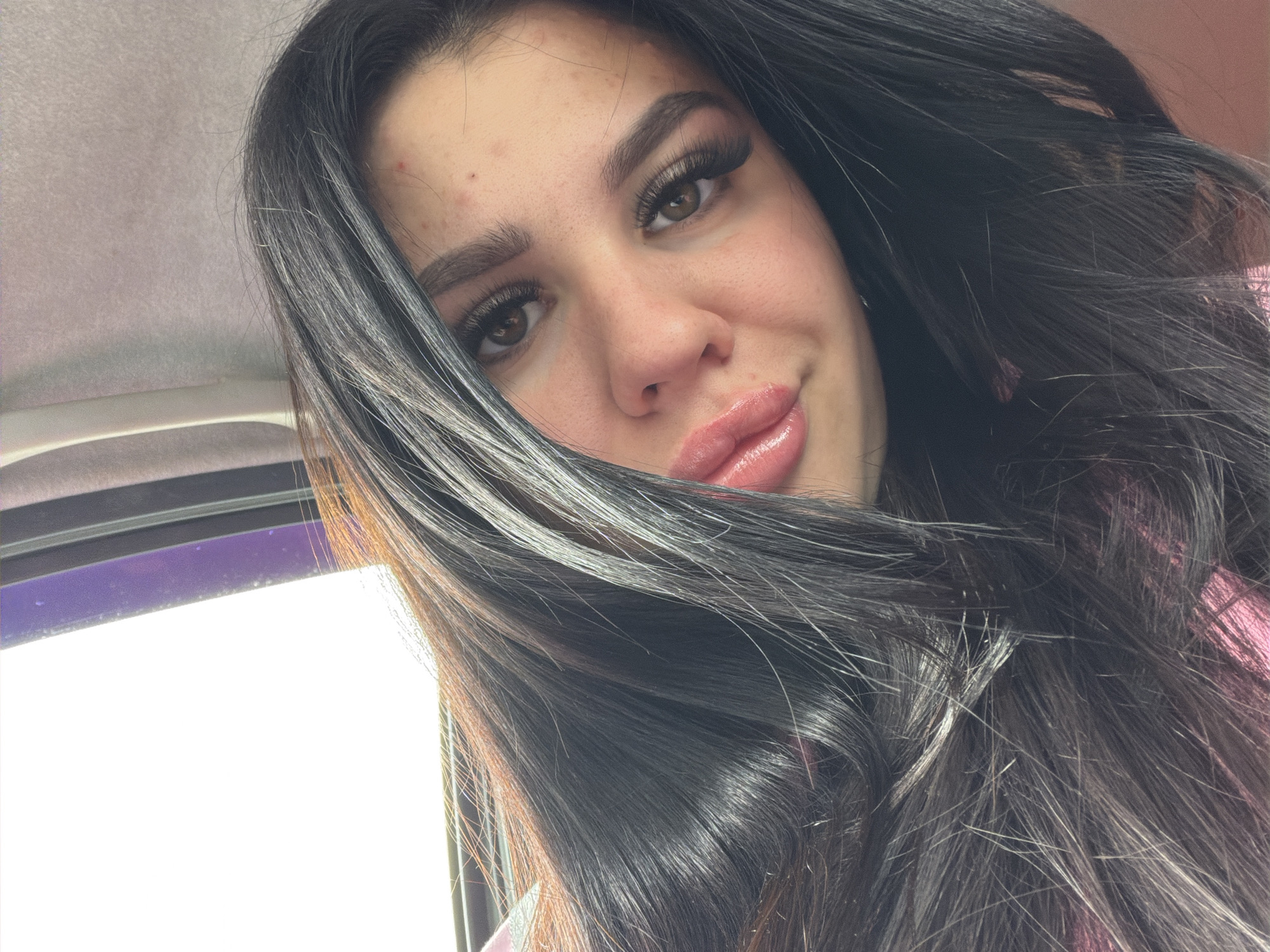 A close-up selfie photograph shows a woman with dark hair and brown eyes, likely inside a vehicle. The presence of a window frame suggests she may be in a car. The background is obscured but appears to be a bright outdoor scene. She is wearing artificial eyelashes and has a few blemishes on her forehead.
There is no verifiable information to identify any specific location in the city of Фоки, Russia, within the context of this image.